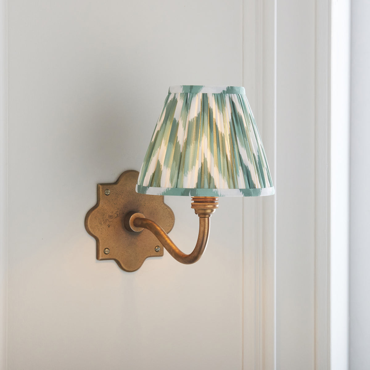 Endon Anvil Curve antique brass wall light lit against pale grey panelled walls, Cotswold green zigzag shade on a curved brass arm