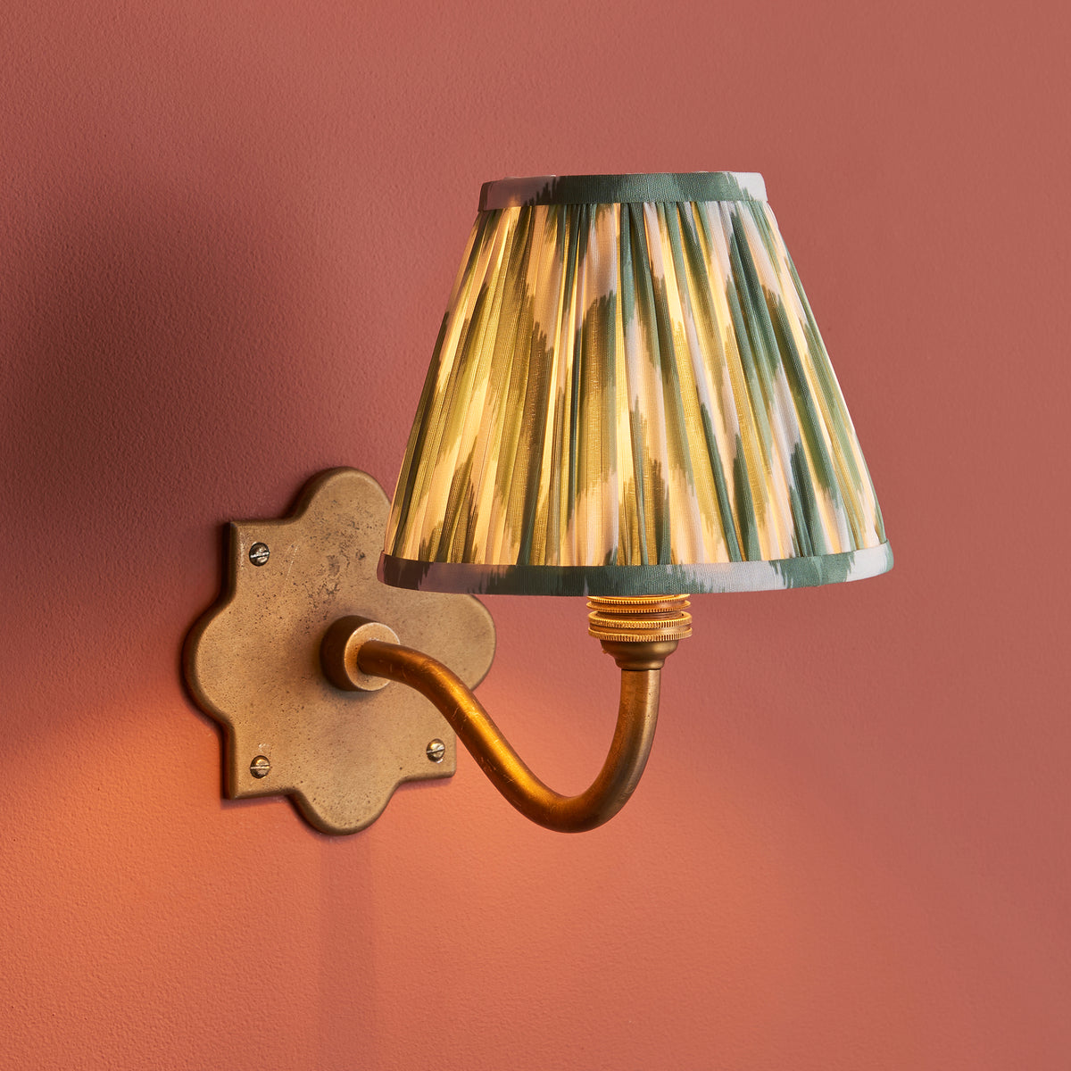 Endon Anvil Curve wall light lit on a terracotta pink wall, antique brass backplate and arm with a glowing green zigzag pleated shade