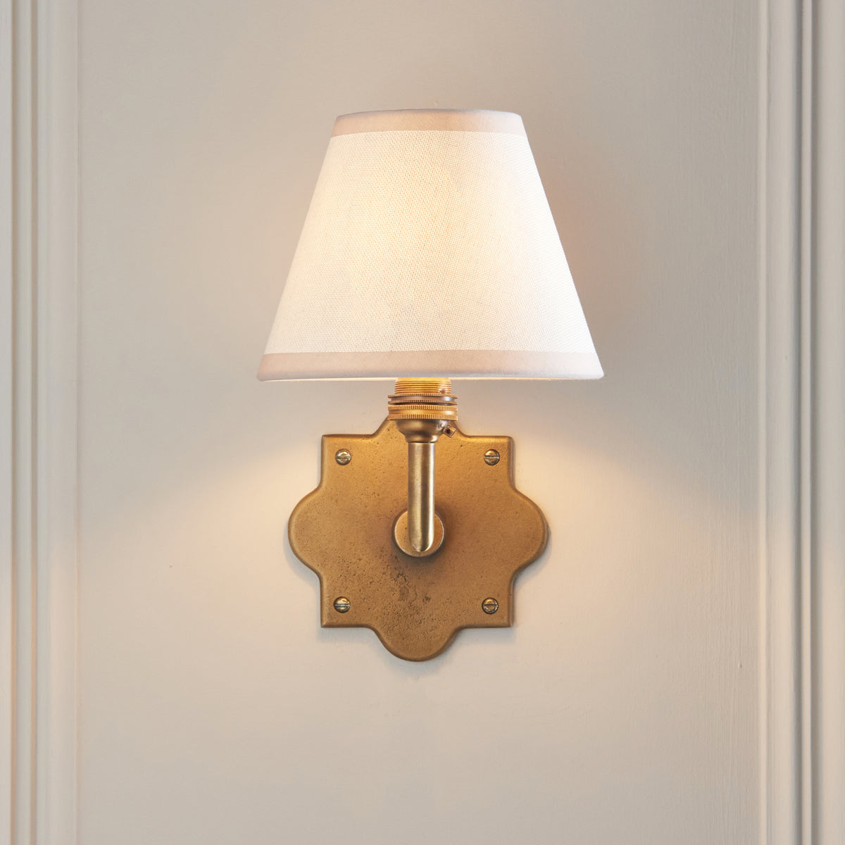 Endon Anvil Angle wall light lit on a cream panelled wall, quatrefoil antique brass backplate centred beneath the vintage white shade