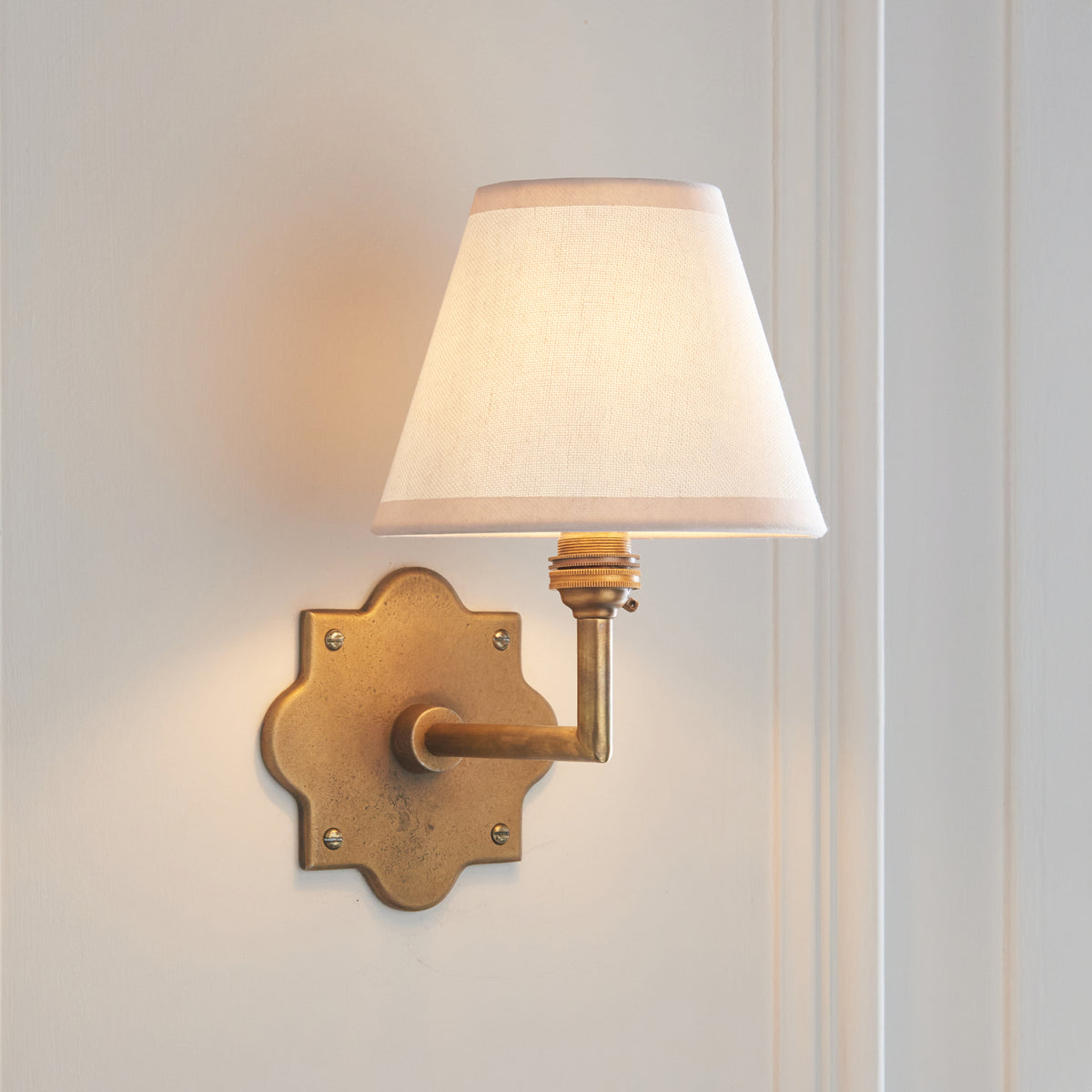 Endon Anvil Angle antique brass wall light lit on a cream panelled wall, backplate to the left of the vintage white Ivy shade