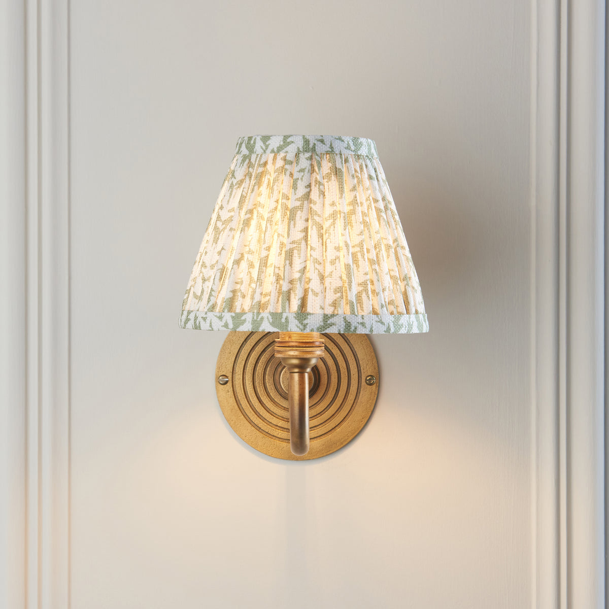 Endon Ripple Curve wall light lit on a panelled wall, green leaf pleated shade sitting directly above the ringed antique brass backplate