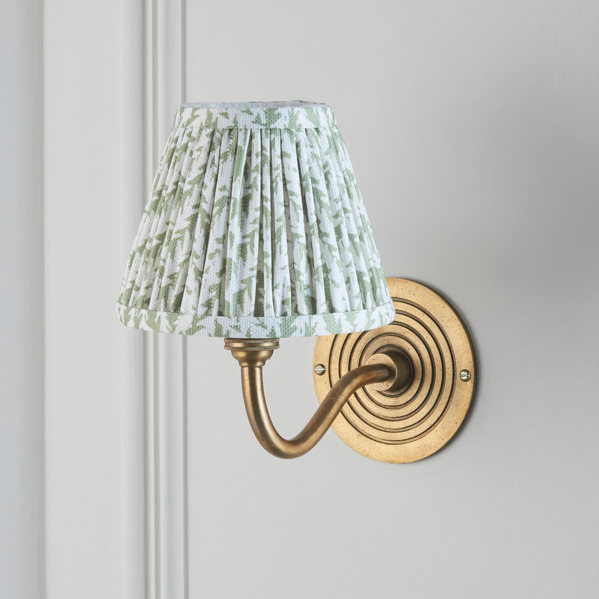 Endon Ripple Curve wall light switched off on a pale panelled wall, showing the sage green leaf print shade and antique brass ringed backplate