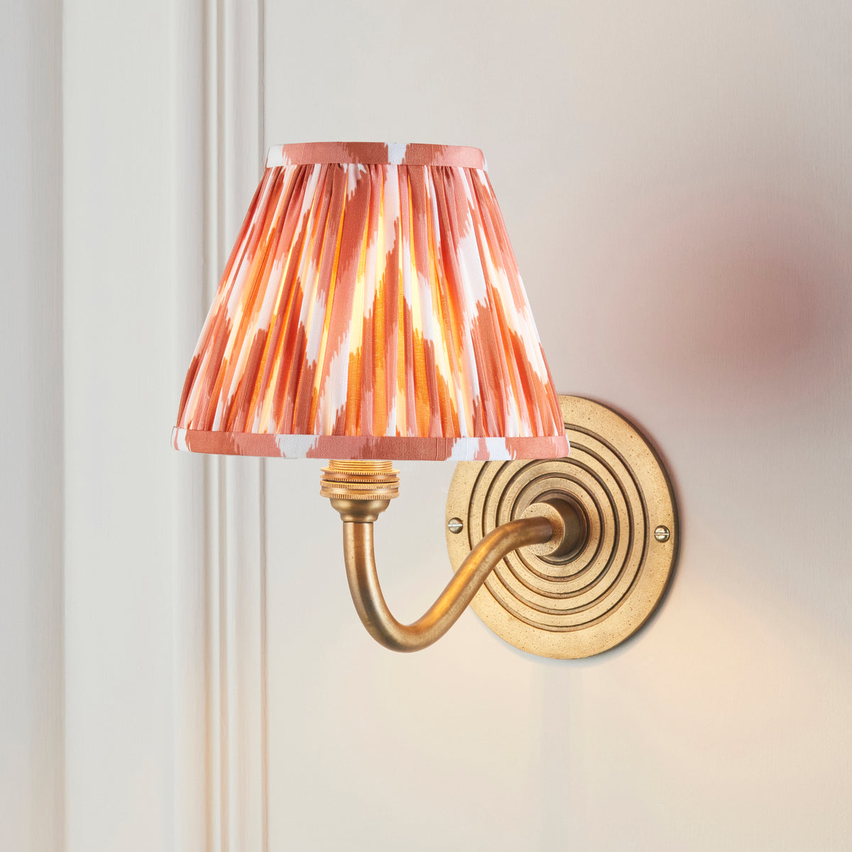 Endon Ripple Curve wall light lit on a cream panelled wall, apricot orange zigzag shade glowing above the antique brass ringed backplate