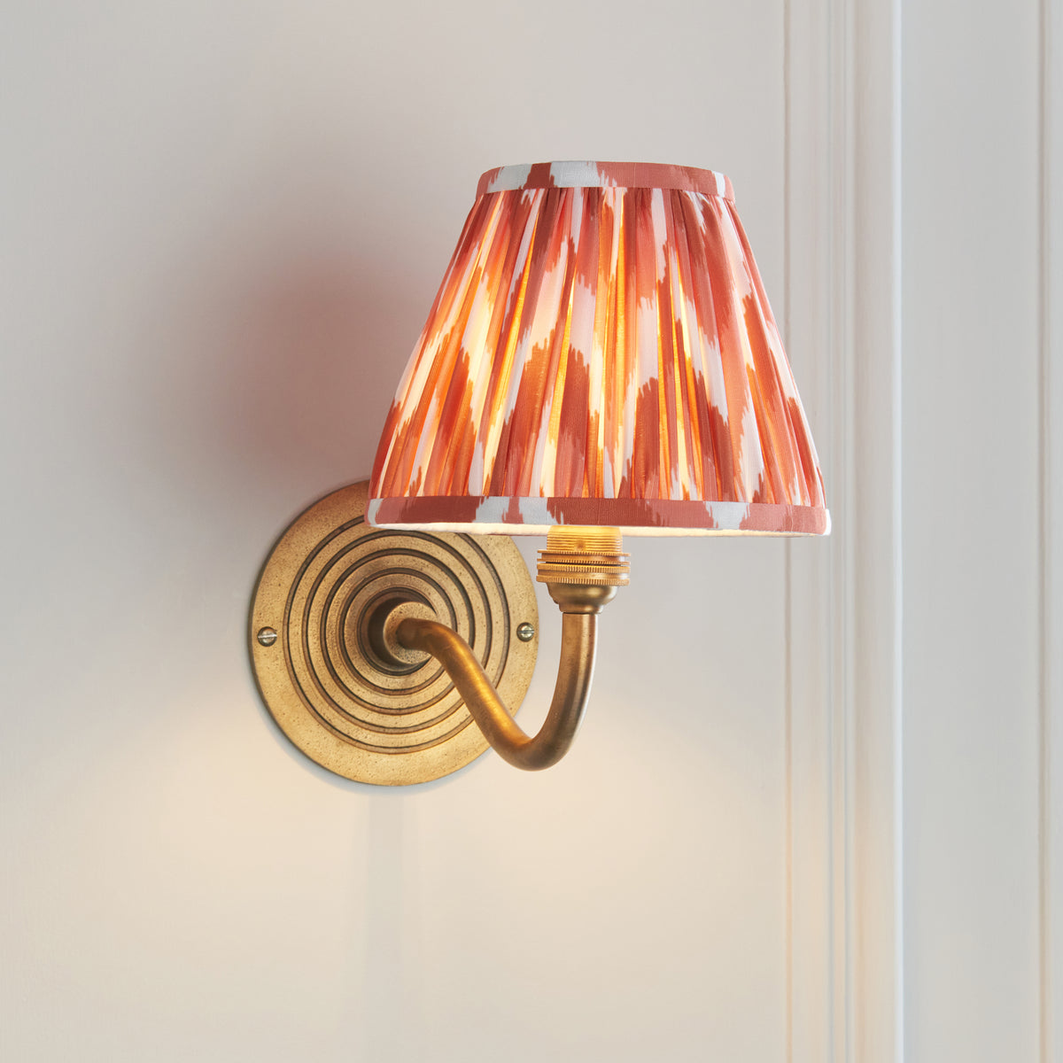 Endon Ripple Curve antique brass wall light lit in a cream panelled room, curved arm and brass lamp holder visible beneath the apricot orange zigzag shade