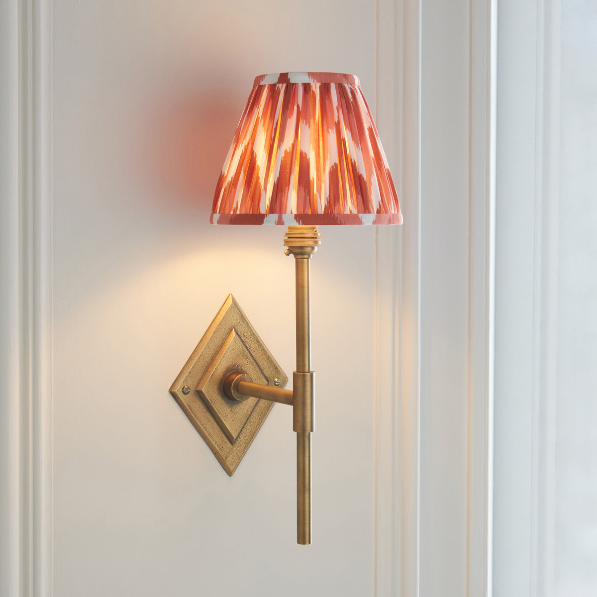 Endon Diamond Tall wall light lit on a pale panelled wall beside a window, antique brass with apricot orange zigzag pleated shade