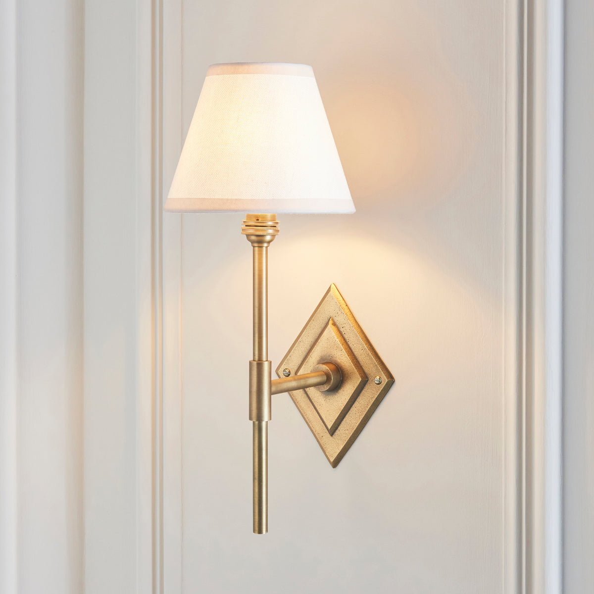 Endon Diamond Tall antique brass wall light lit on a cream panelled wall, vintage white shade glowing above the diamond backplate