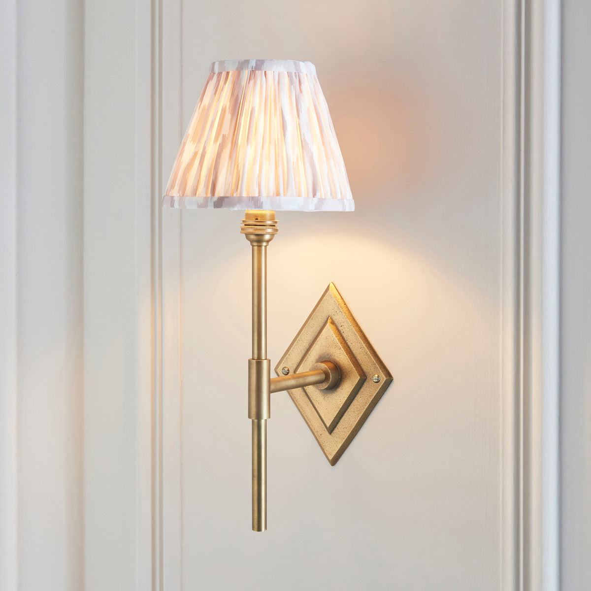 Endon Diamond Tall wall light lit on a cream panelled wall, antique brass arm with the pink and cream ikat shade casting a warm glow