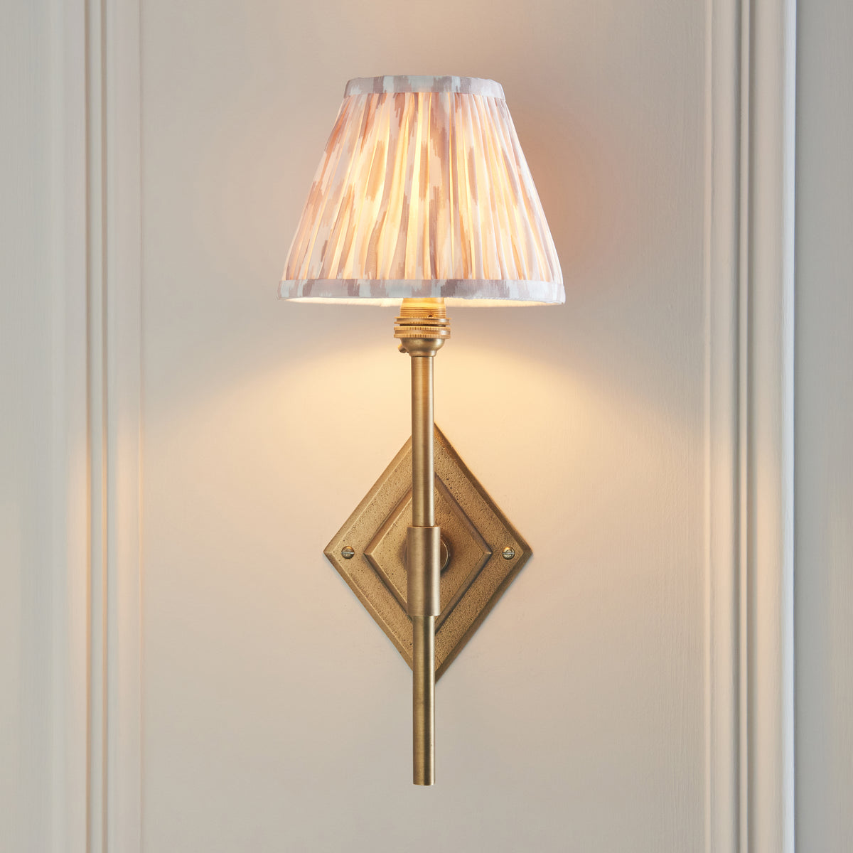 Endon Diamond Tall wall light lit on a cream panelled wall, the antique brass diamond backplate visible behind the slim stem and ikat shade