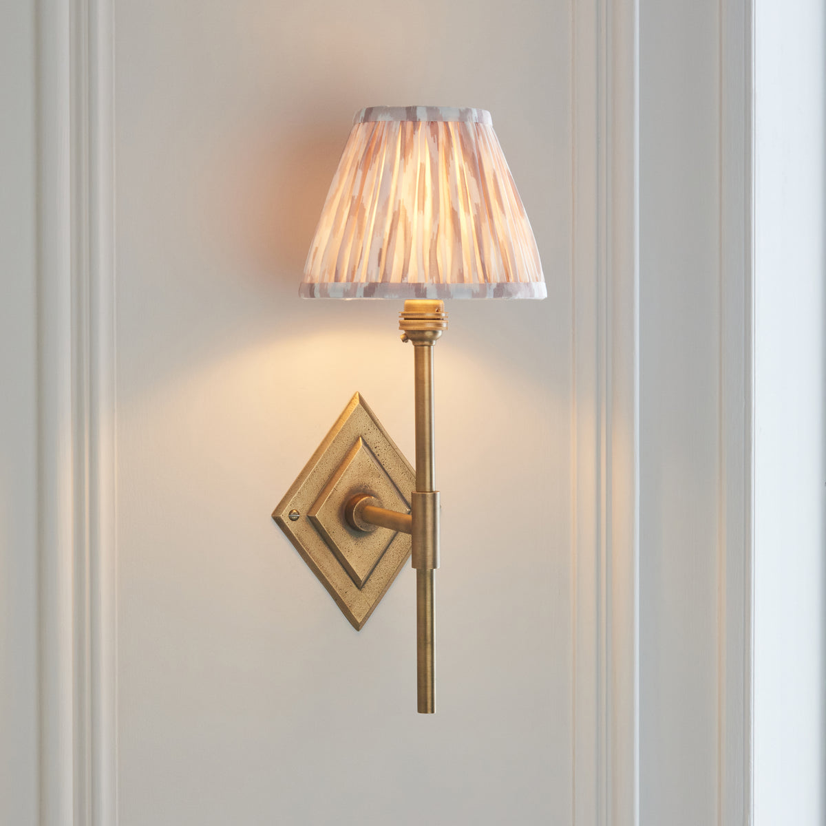 Endon Diamond Tall antique brass wall light lit on a pale panelled wall, the neutral ikat pleated shade glowing above the stem