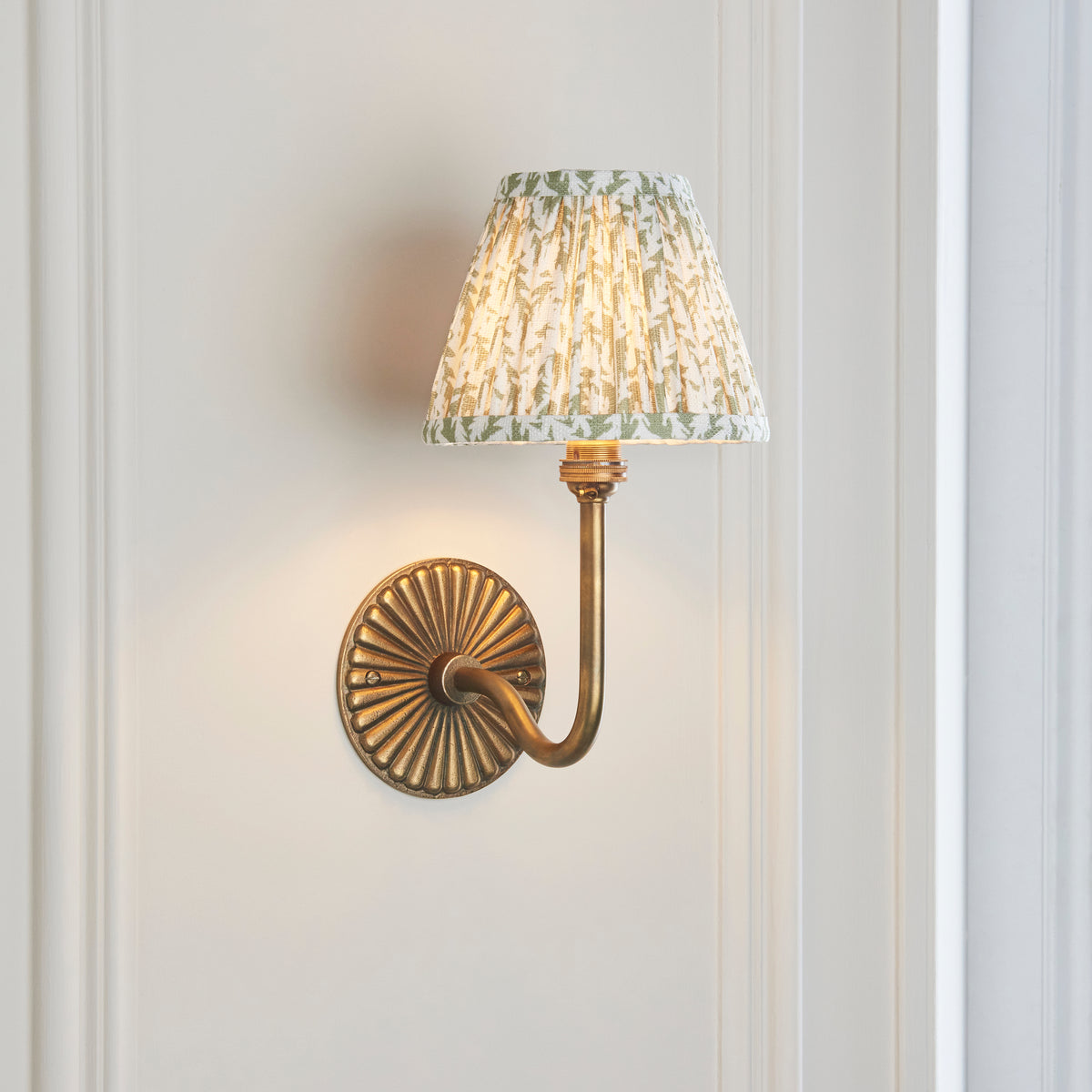 Endon Dahlia Swan wall light with green leaf shade lit on a cream panelled wall, curved brass arm and fluted backplate visible