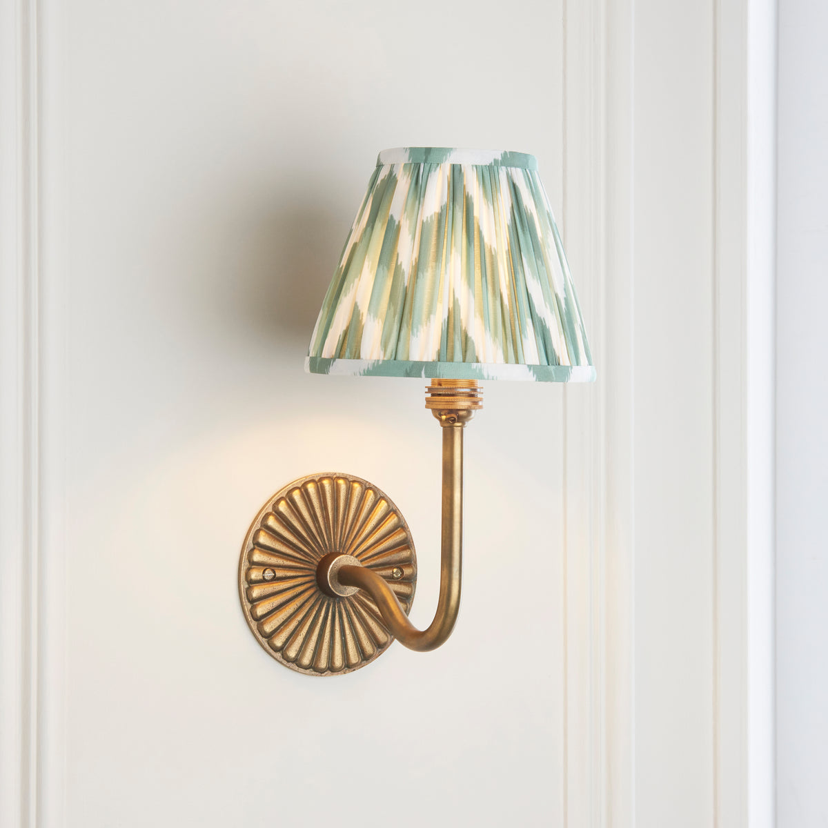 Endon Dahlia Swan antique brass wall light on a cream panelled wall, lit, curved arm and Cotswold green pleated shade