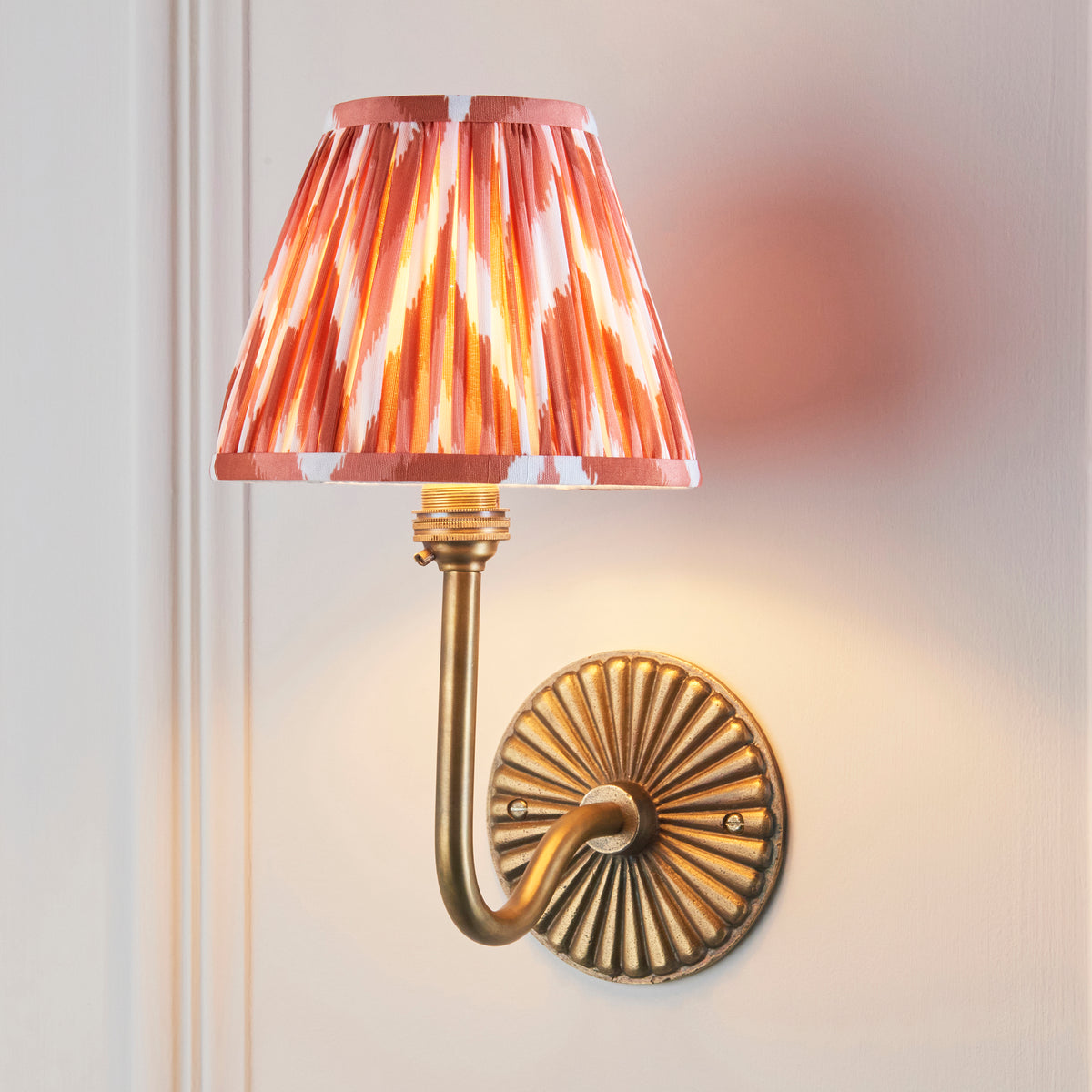 Endon Dahlia Swan wall light lit on a cream panelled wall, apricot orange zigzag shade glowing above the antique brass sunburst backplate