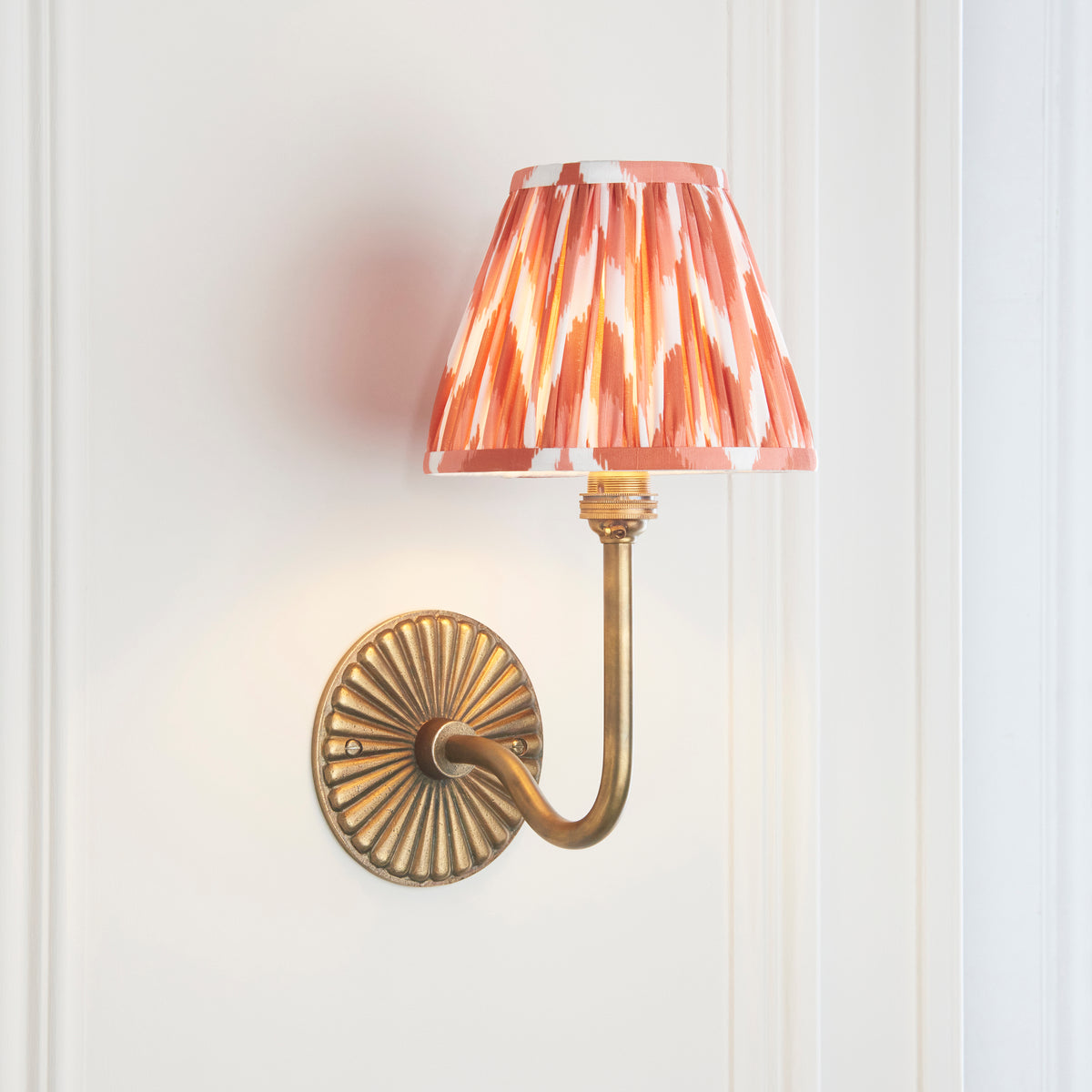 Endon Dahlia Swan wall light lit on a cream panelled wall showing the curved antique brass arm and apricot orange zigzag shade