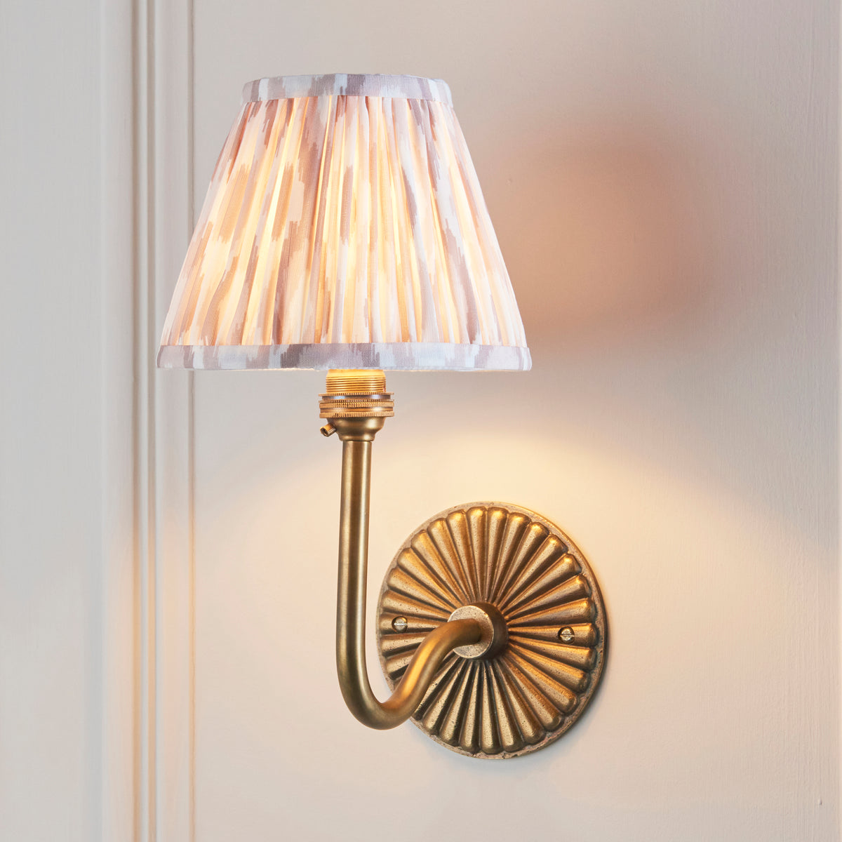 Endon Dahlia Swan antique brass wall light lit on a cream panelled wall, the ikat shade glowing warm pink and cream