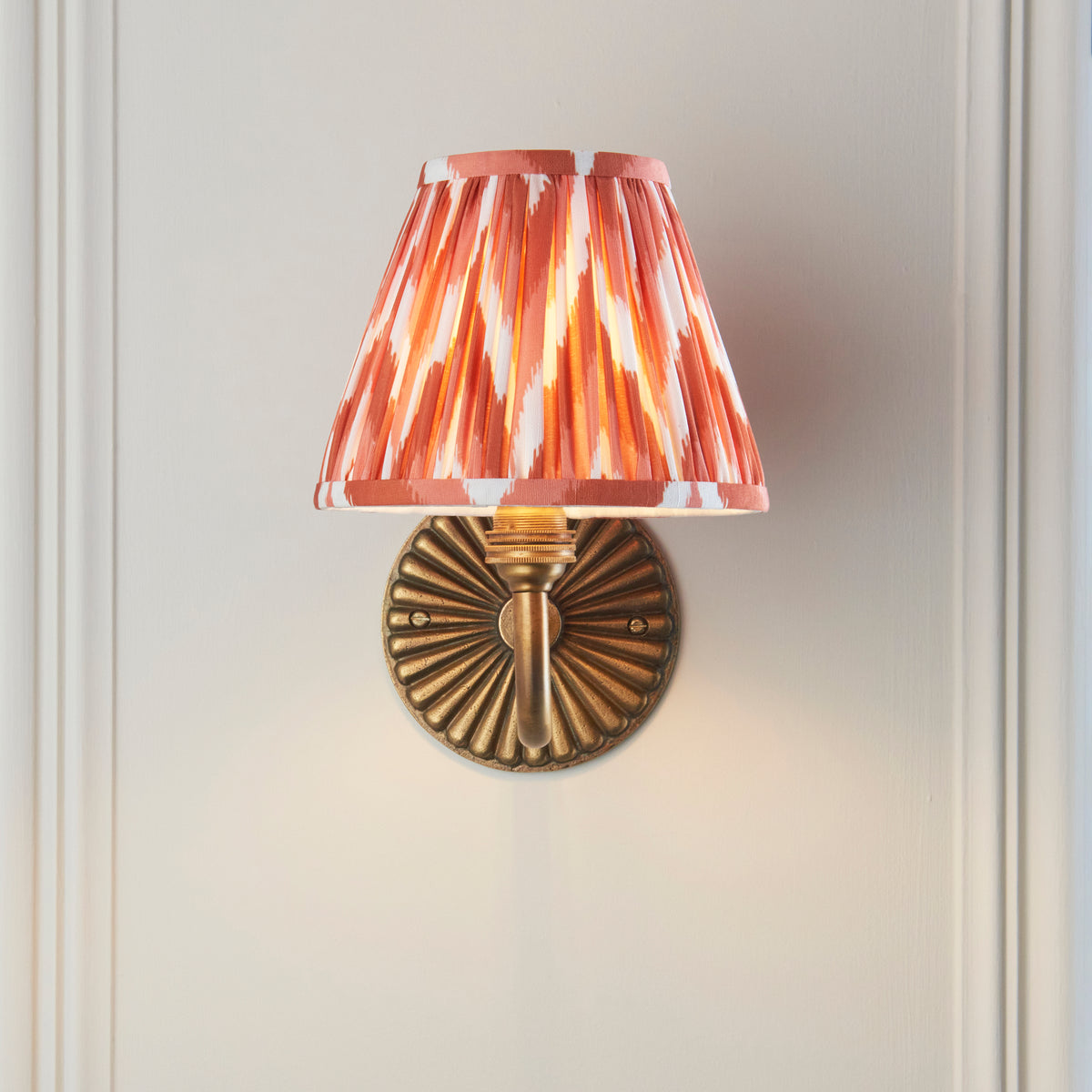 Endon Dahlia Curve wall light lit on a cream panelled wall, apricot orange zigzag shade sitting above the fluted brass backplate