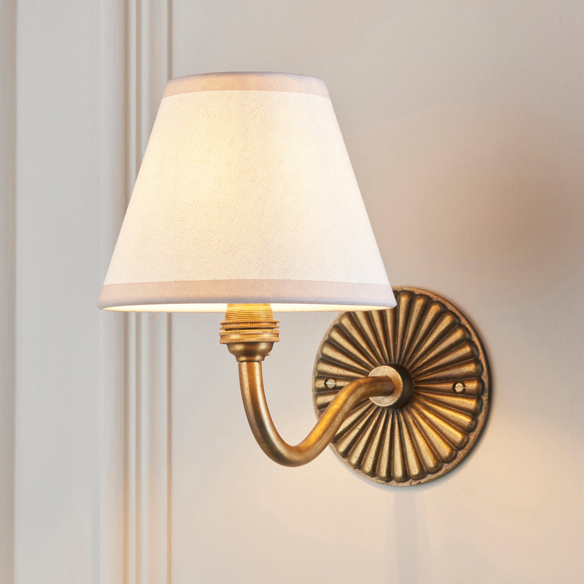 Endon Dahlia Curve antique brass wall light lit on a cream panelled wall, vintage white shade glowing above the fluted backplate
