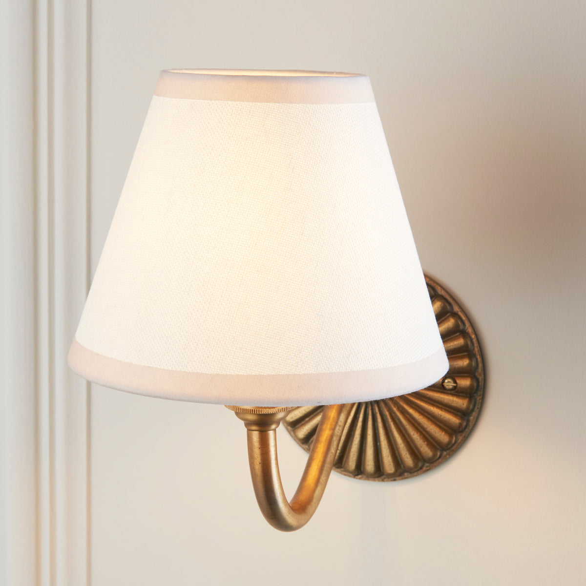 Close up of the Endon Dahlia Curve wall light lit, vintage white fabric shade glowing with the brass arm and fluted backplate behind