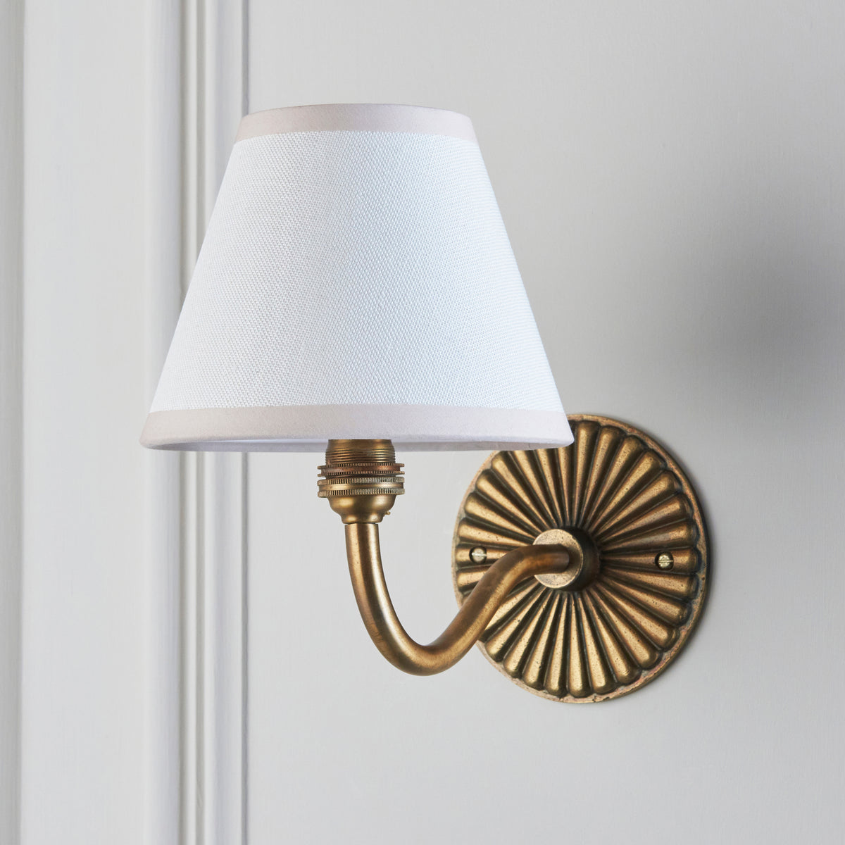 Endon Dahlia Curve wall light unlit on a pale grey panelled wall, fluted antique brass backplate, curved arm and white Ivy shade