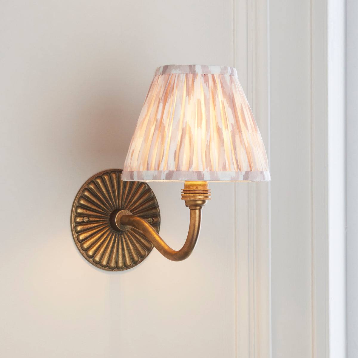 Endon Dahlia Curve antique brass wall light glowing on a pale panelled wall, swan neck arm and pink ikat shade