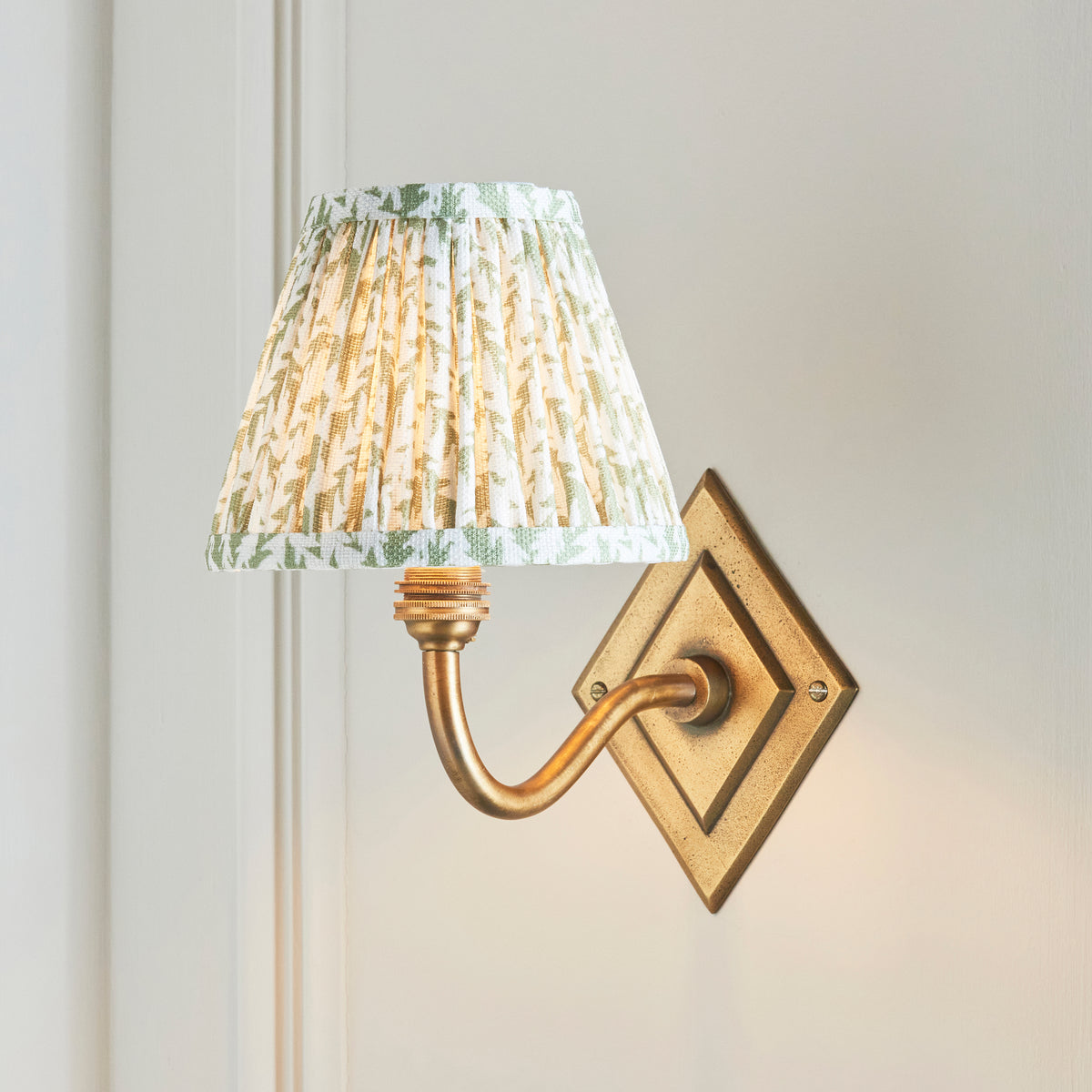 Endon Diamond Curve antique brass wall light lit on a cream panelled wall, green leaf pleated shade held out on the curved arm