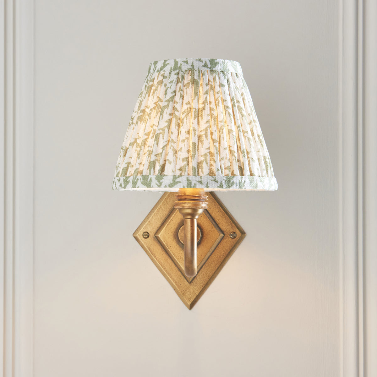 Lit Endon Diamond Curve wall light on a cream panelled wall, diamond antique brass backplate sitting below the green leaf print shade