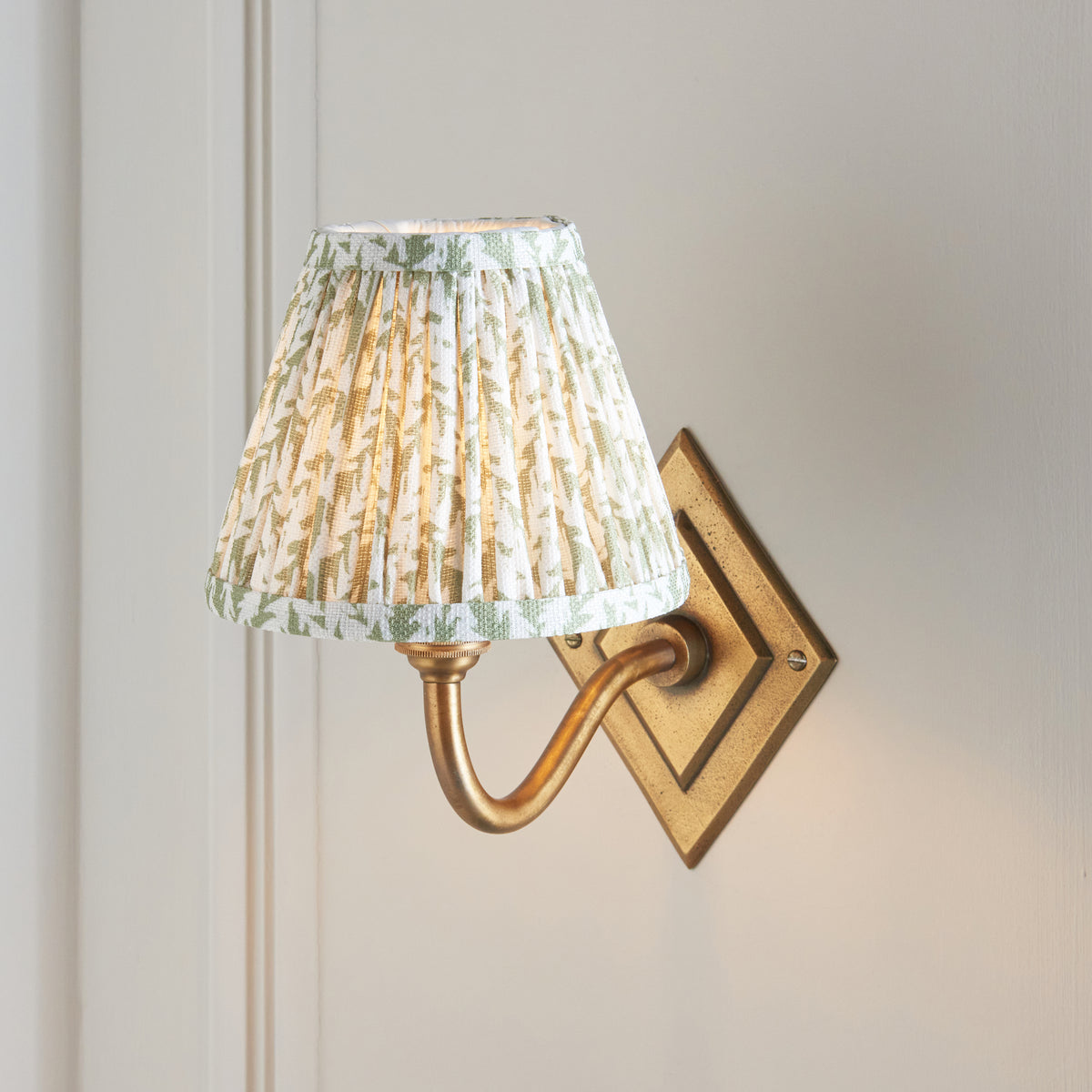 Endon Diamond Curve wall light with a softly lit green leaf pleated shade and antique brass swan neck arm on a cream panelled wall