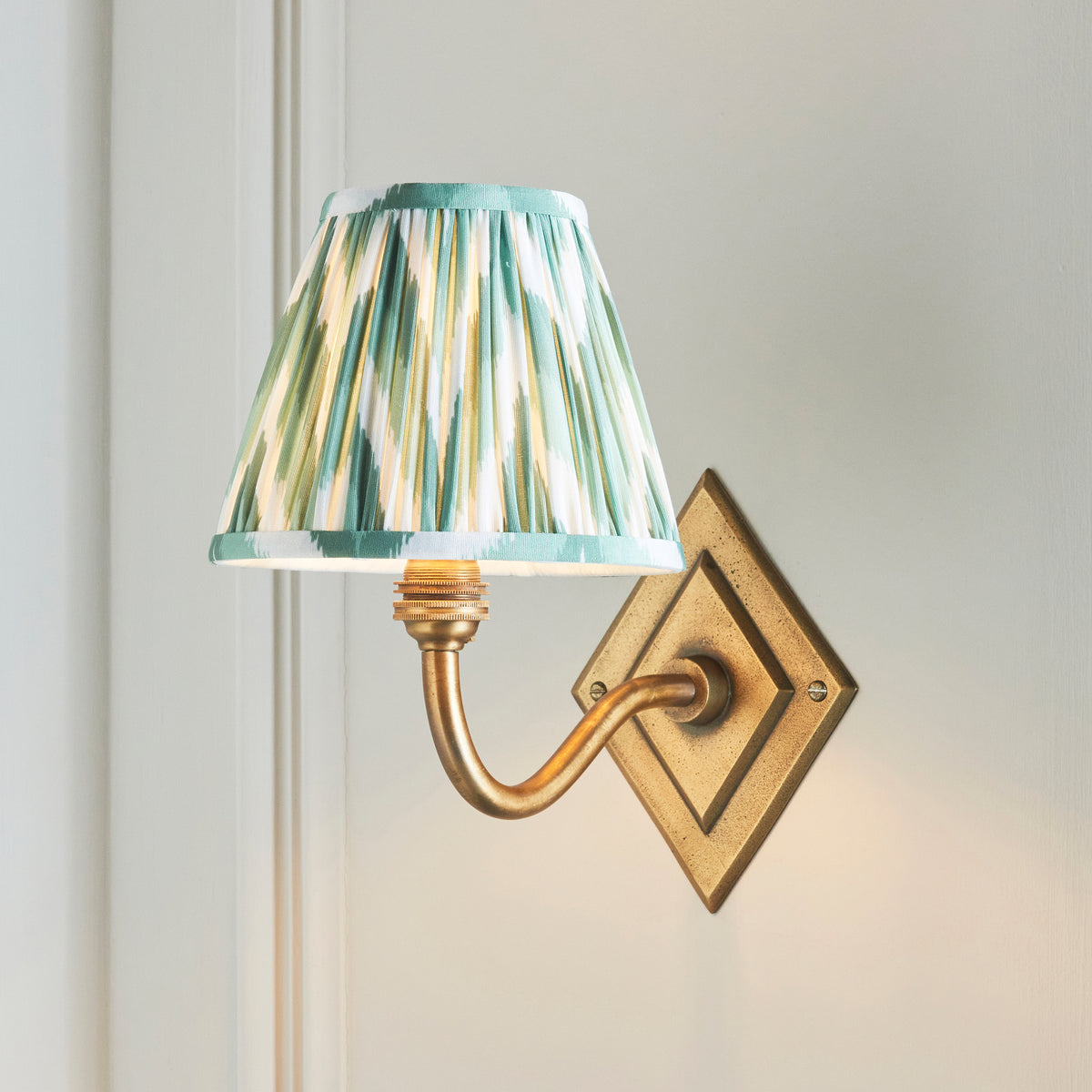 Endon Diamond Curve wall light lit on a cream panelled wall, green and white zigzag shade angled to the left of the diamond brass backplate