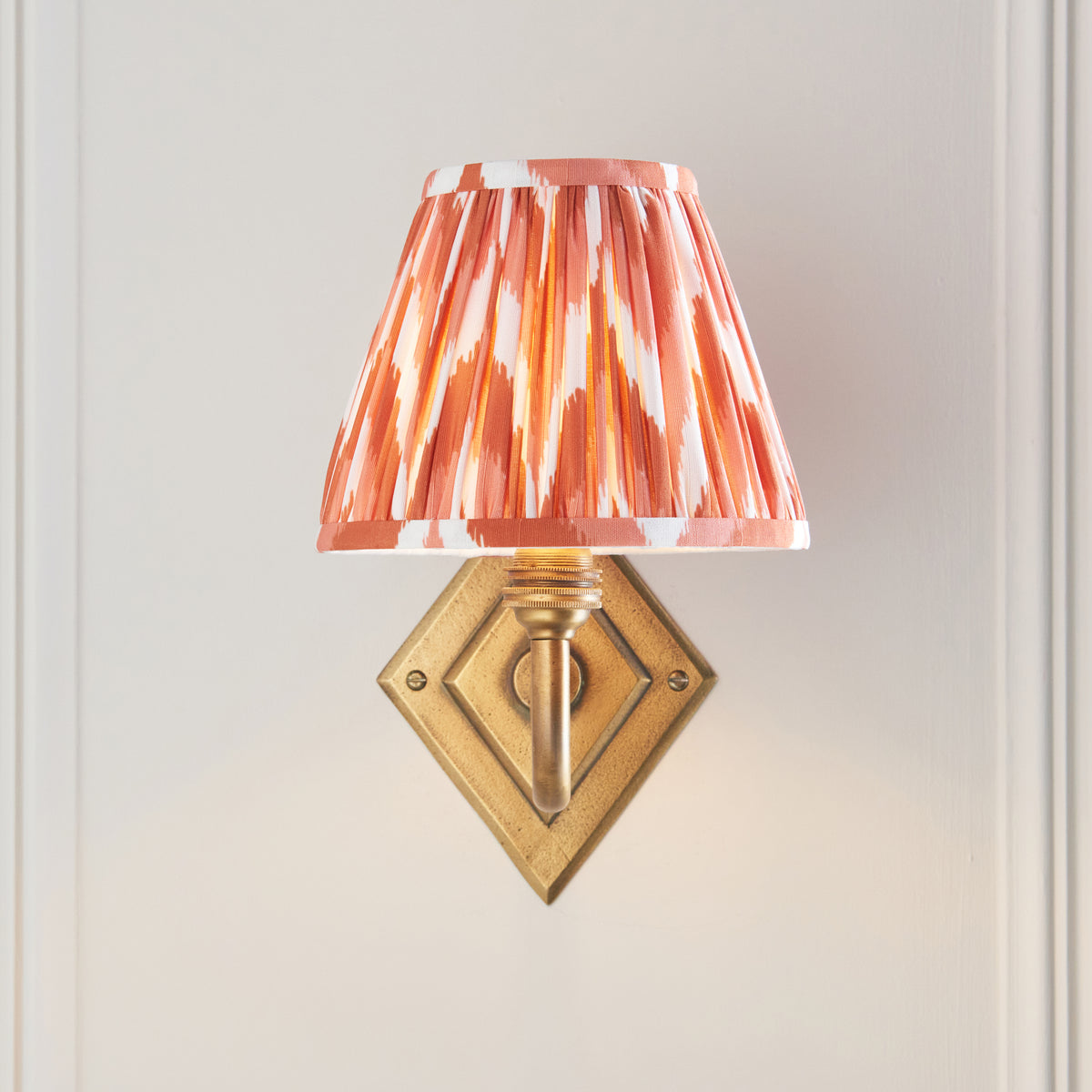 Endon Diamond Curve wall light lit on a cream panelled wall, diamond brass backplate fully visible beneath the apricot orange zigzag shade