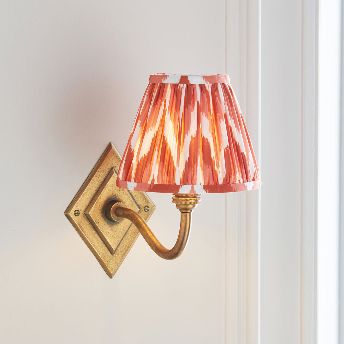 Endon Diamond Curve antique brass wall light lit in a cream panelled room, apricot orange zigzag shade with the curved arm and diamond backplate showing