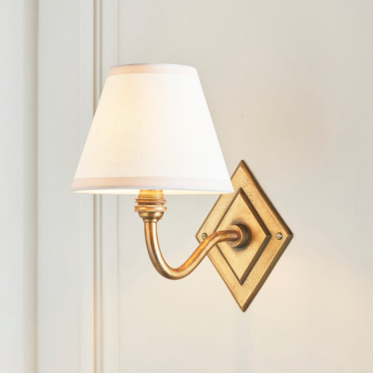 Endon Diamond Curve antique brass wall light with white Ivy shade, lit, mounted on a cream panelled wall