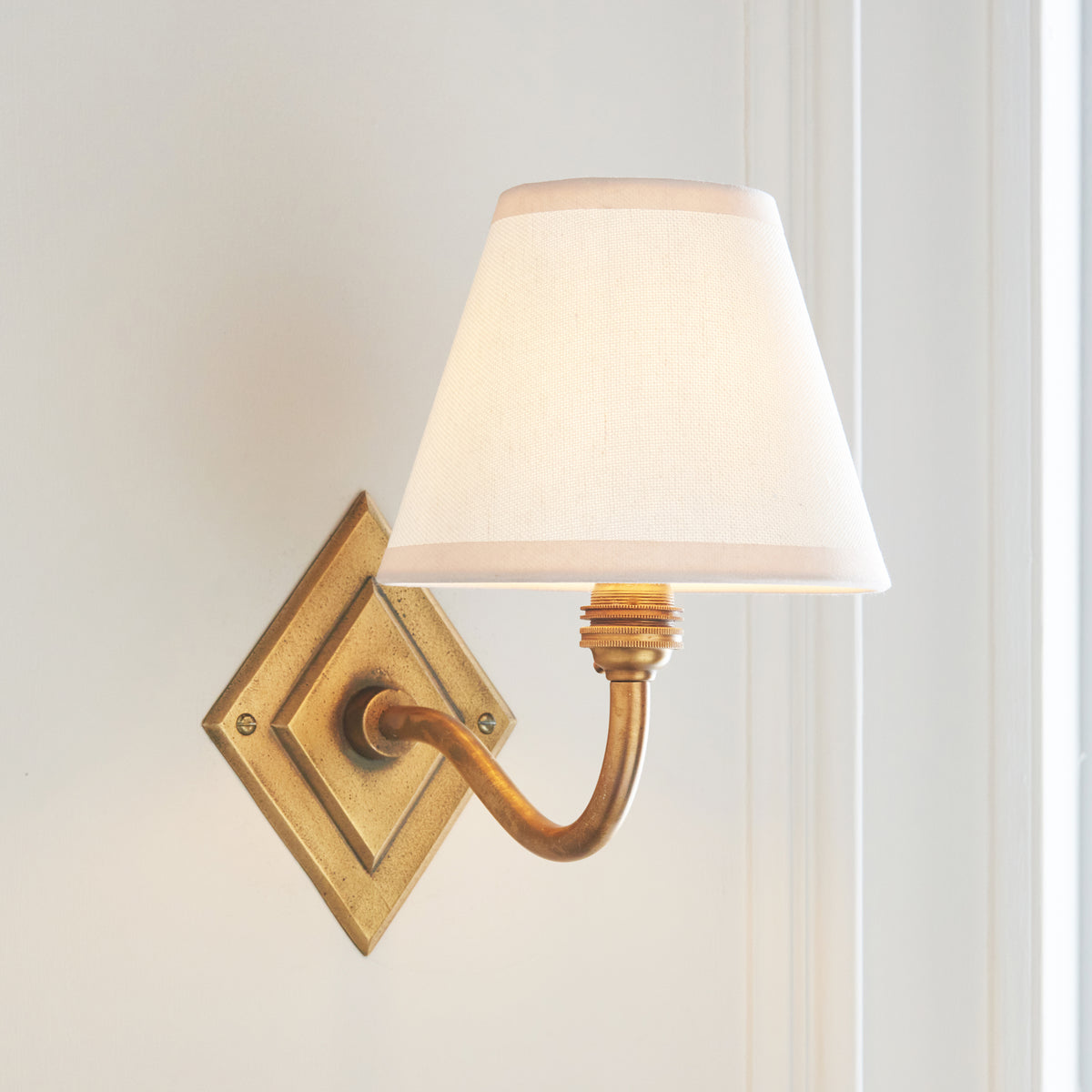 Endon Diamond Curve antique brass wall light lit, white Ivy shade glowing, curved arm visible against a pale panelled wall