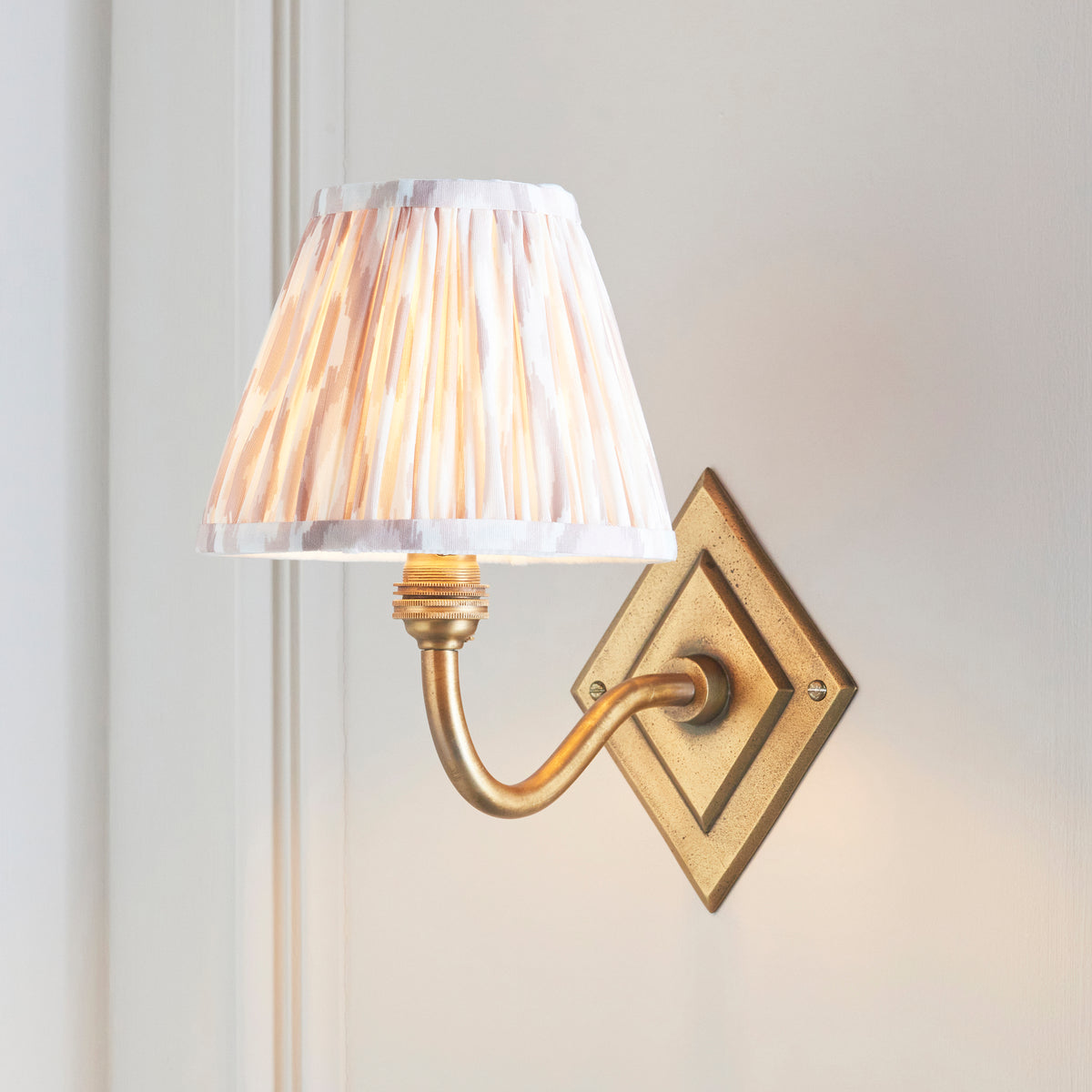 Endon Diamond Curve antique brass wall light switched on, blush ikat pleated shade glowing against a cream panelled wall