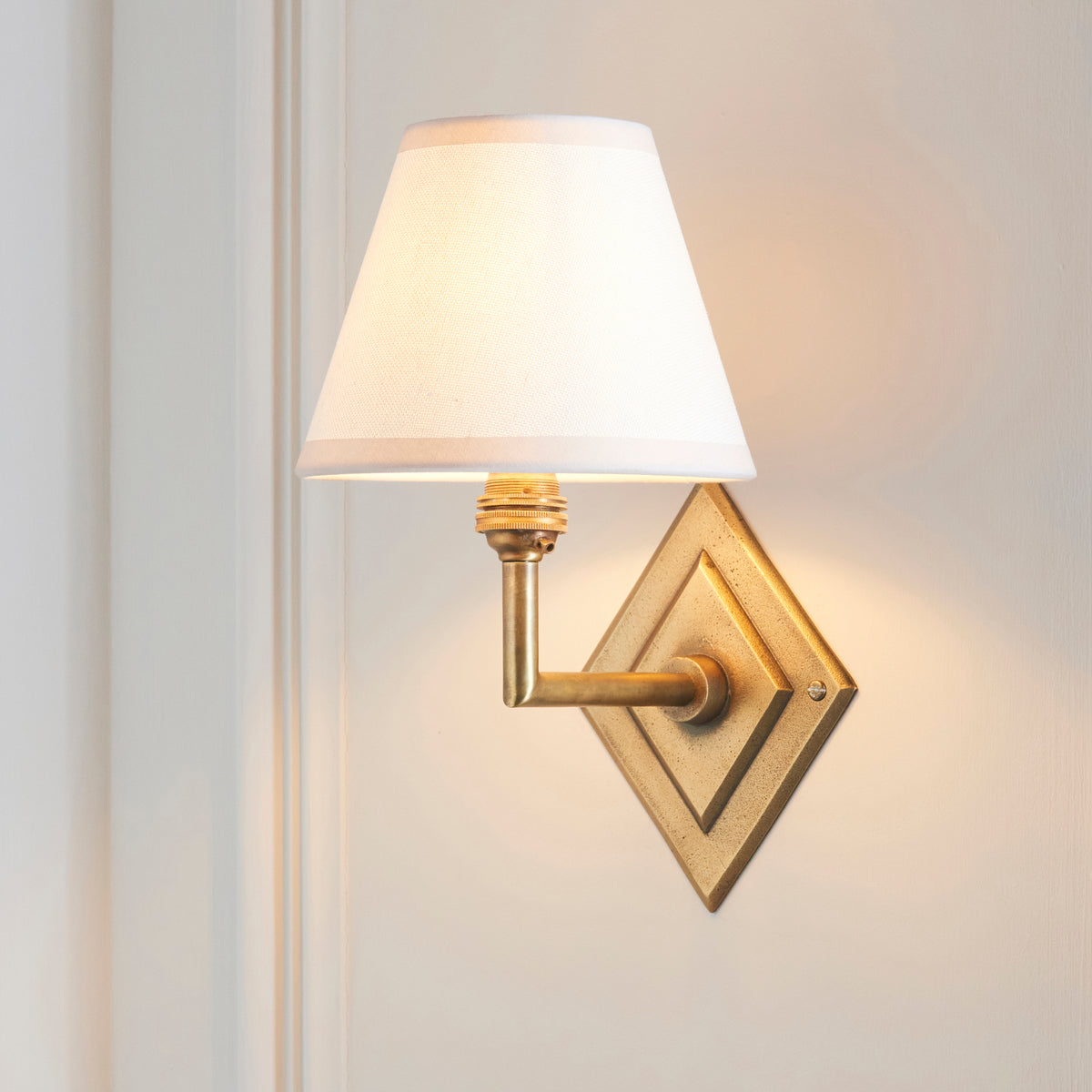 Endon Diamond Angle antique brass wall light switched on, white Ivy shade glowing against an off white panelled wall
