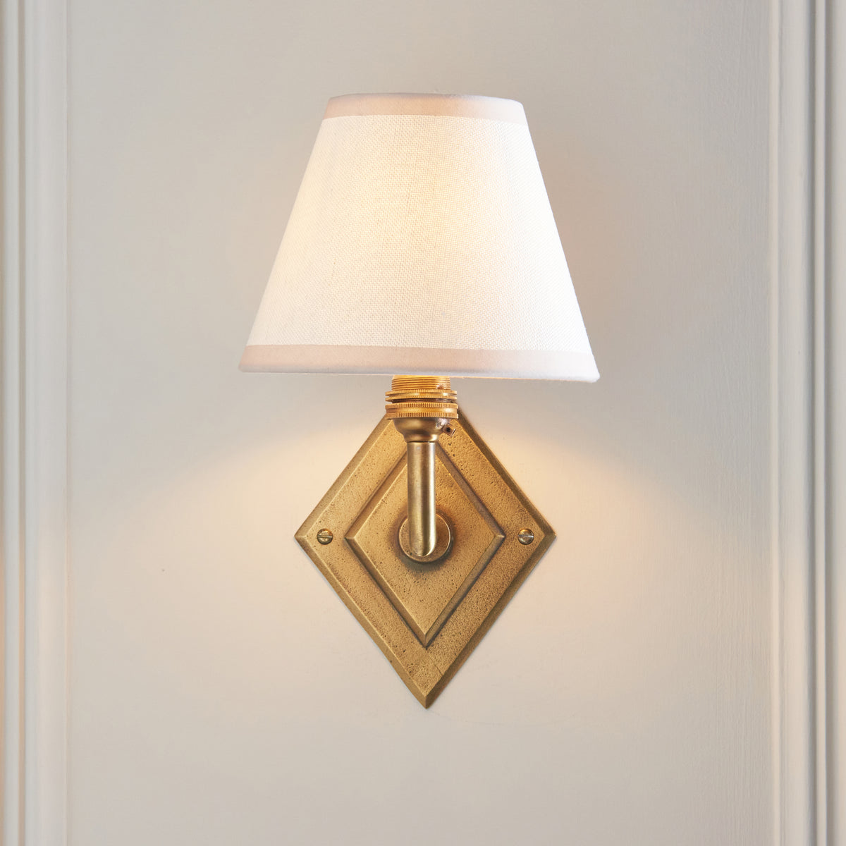 Endon Diamond Angle wall light lit on a cream panelled wall, stepped diamond brass backplate fully visible below the white shade
