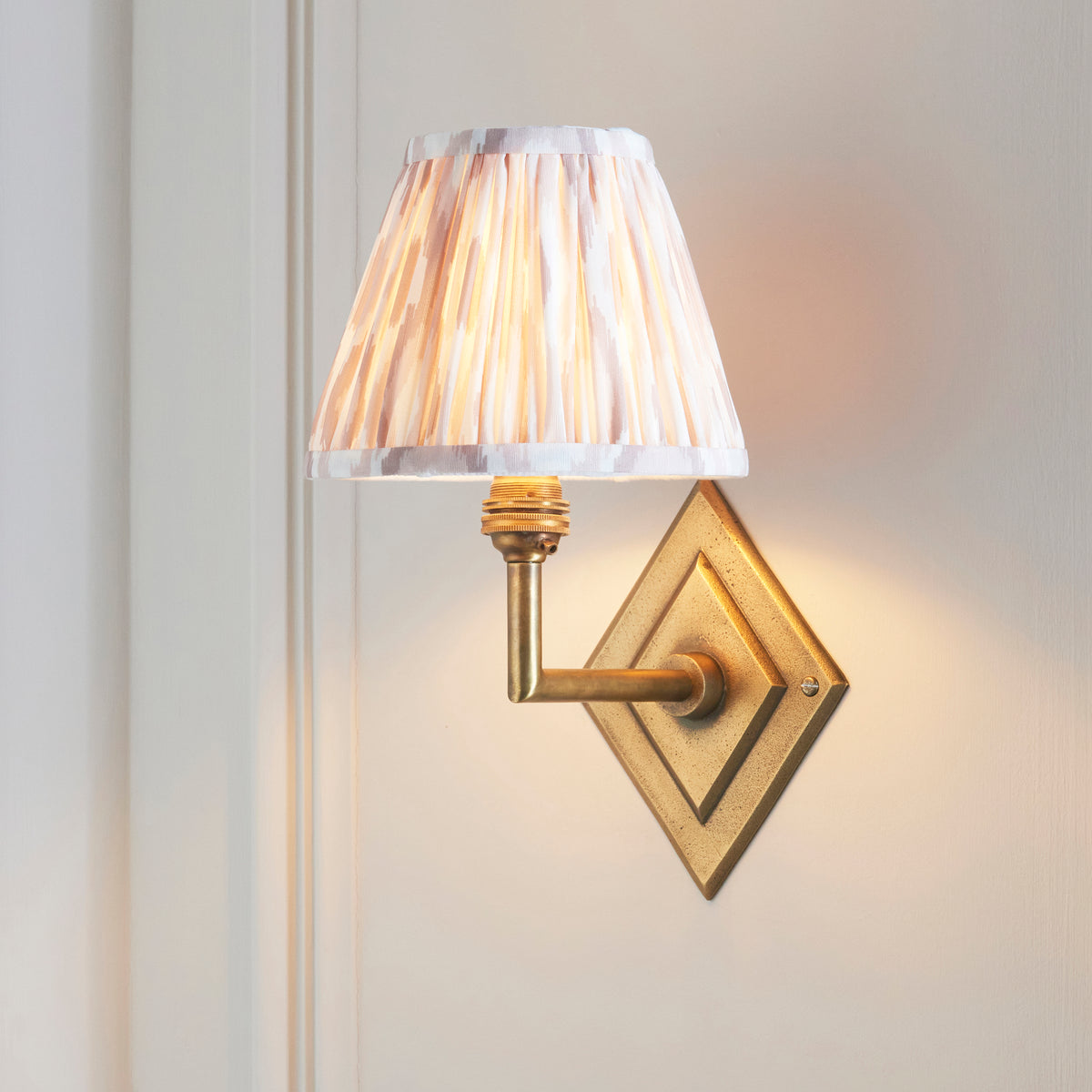Endon Diamond Angle wall light switched on against a cream panelled wall, antique brass backplate and lit pink and beige ikat pleated shade