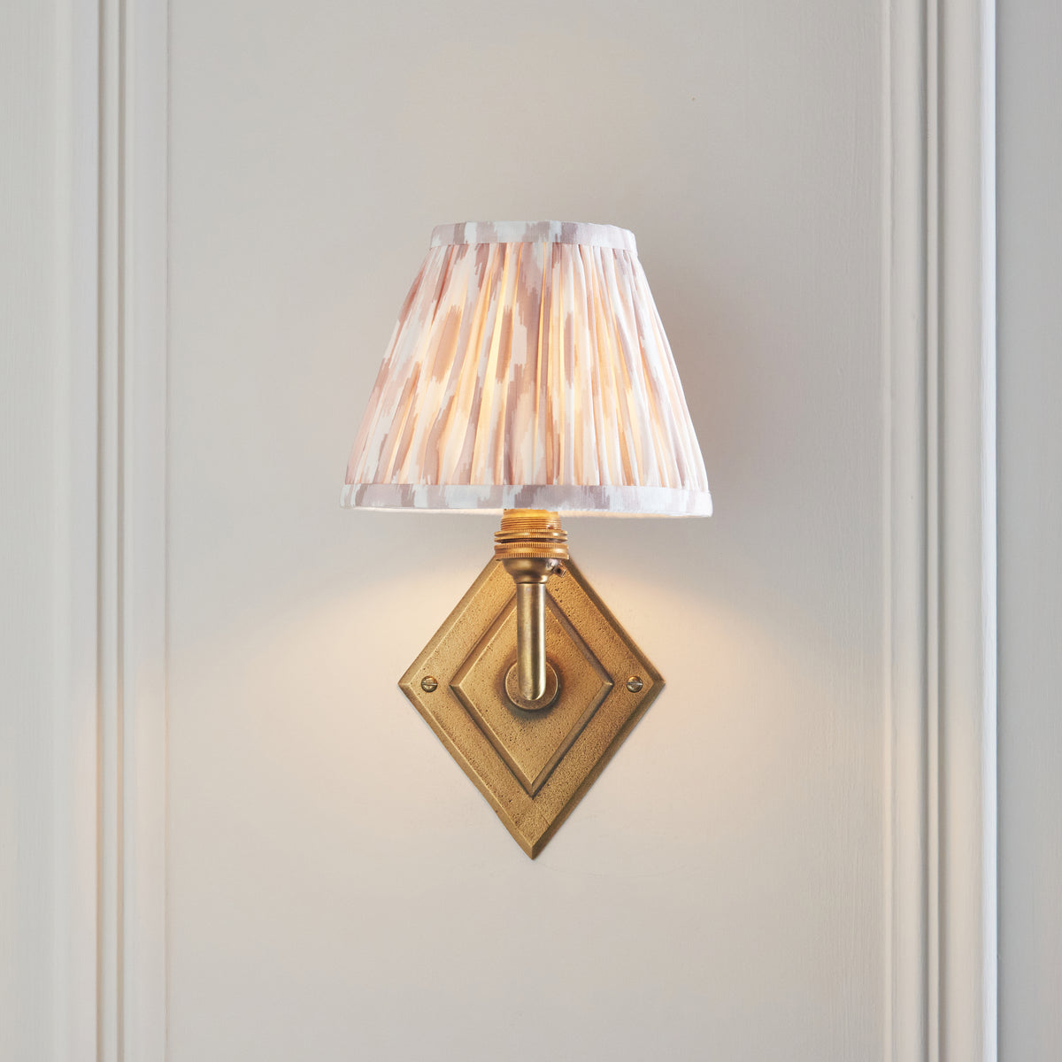 Endon Diamond Angle antique brass wall light lit on a cream panelled wall, arm reaching upward to the glowing neutral ikat shade