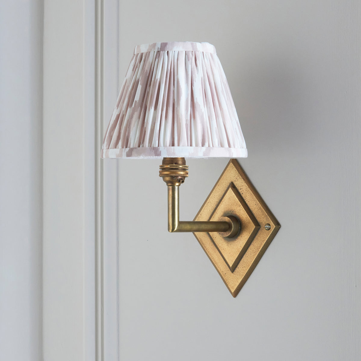 Endon Diamond Angle wall light switched off on a pale grey panelled wall, antique brass backplate with a pink and white ikat pleated shade