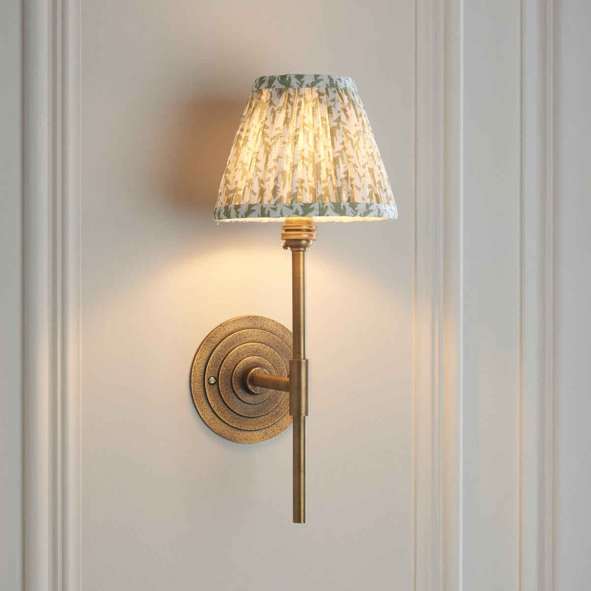 Endon Halo Tall brass wall light switched on against cream wall panelling, pleated green leaf shade and stepped round backplate