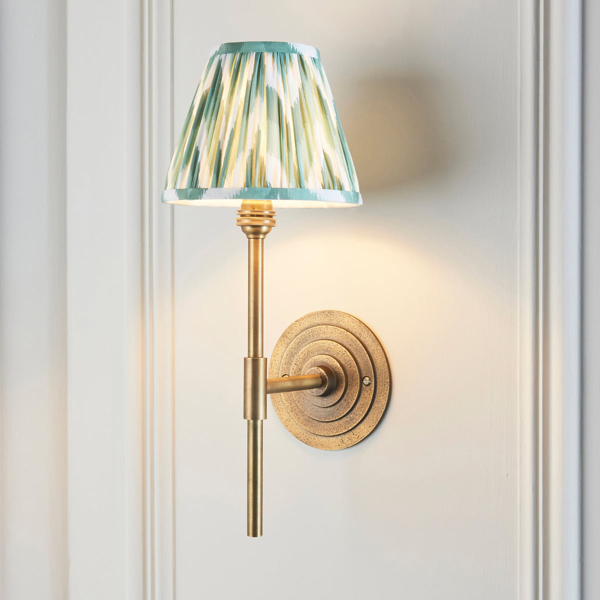 Endon Halo Tall antique brass wall light switched on, green zigzag shade glowing on a cream panelled wall, round stepped backplate to one side of the stem