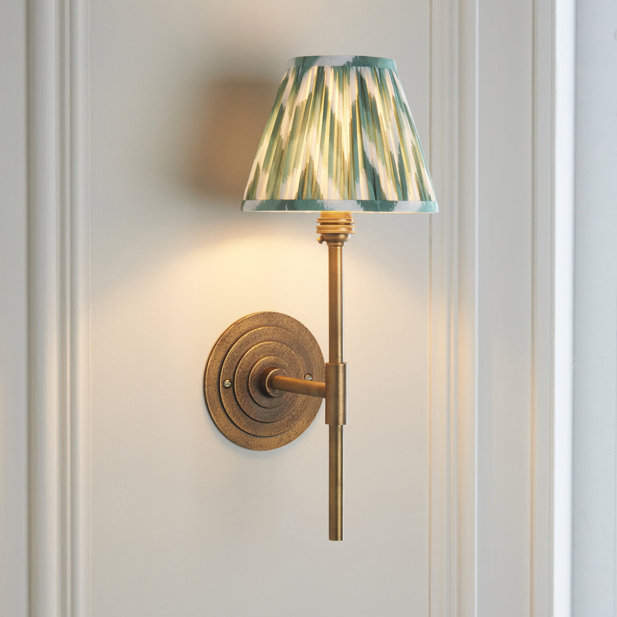 Endon Halo Tall wall light switched on, antique brass stem and green zigzag shade casting warm light up a white panelled wall