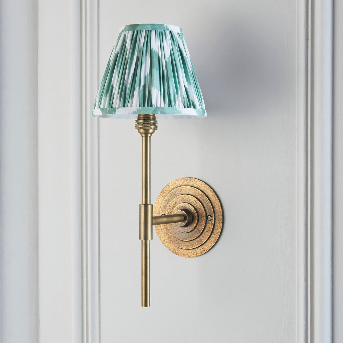 Endon Halo Tall wall light unlit on a pale grey green panelled wall, antique brass halo backplate and green zigzag pleated shade