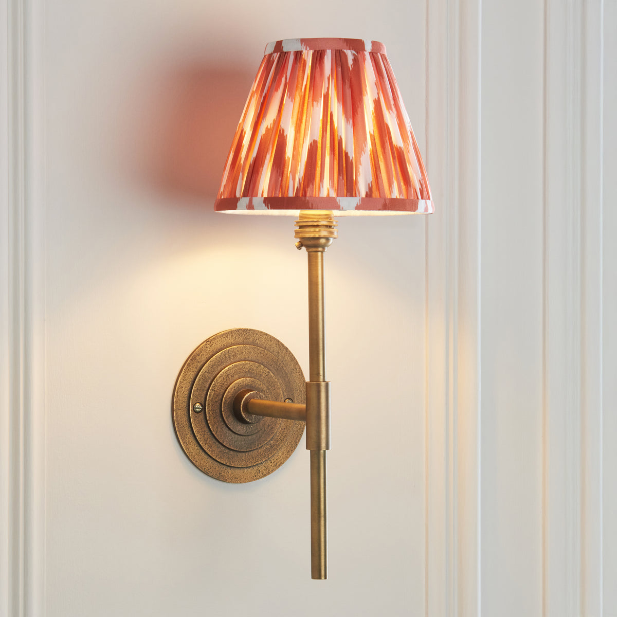 Endon Halo Tall wall light switched on, ringed antique brass backplate and apricot orange pleated shade against cream panelling
