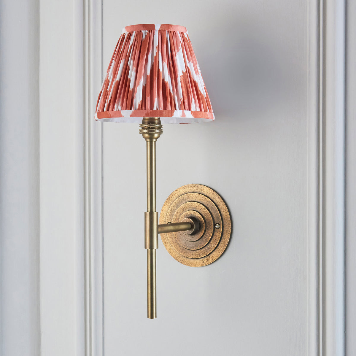 Endon Halo Tall wall light unlit on a pale grey panelled wall, antique brass stem and apricot orange zigzag shade