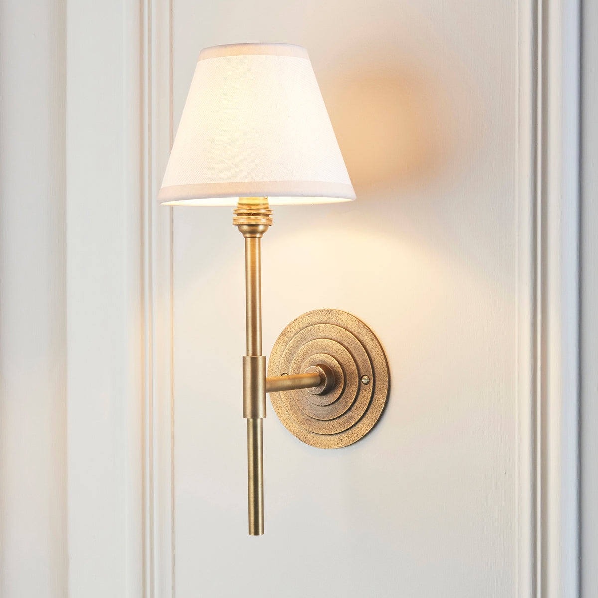 Endon Halo Tall antique brass wall light lit on a cream panelled wall, the vintage white Ivy shade glowing warmly