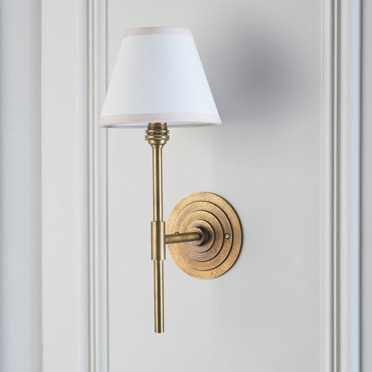 Endon Halo Tall wall light unlit on a pale grey panelled wall, showing the antique brass column and vintage white shade with taped edge