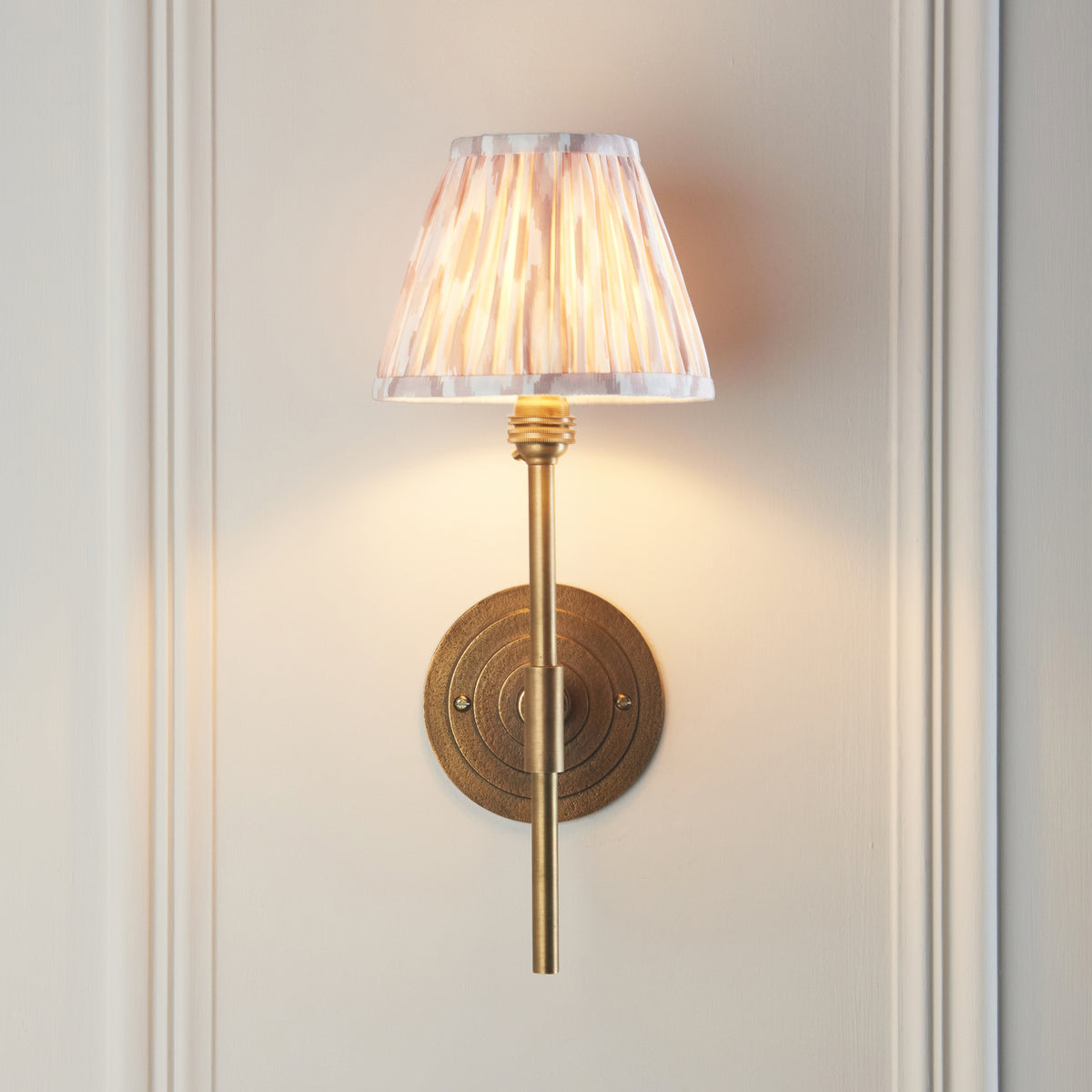 Endon Halo Tall wall light lit on cream panelling, brass stem passing in front of the ringed antique brass backplate, Ikat shade in soft peach and grey