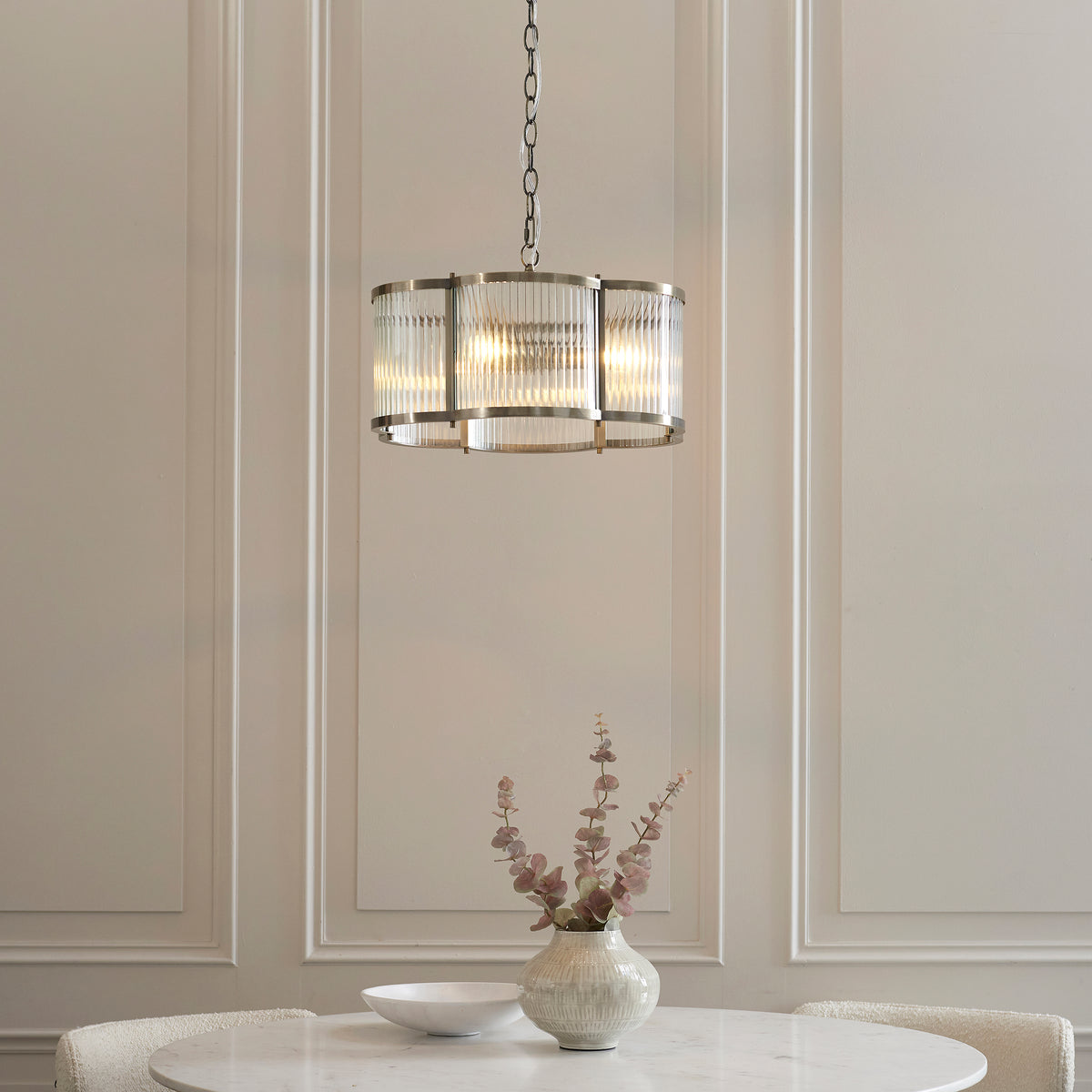Endon Ridgeton Scallop three light pendant with clear ribbed glass panels in a brushed silver tone frame, lit warm above a round dining table with a vase of eucalyptus stems against a wood panelled wall.