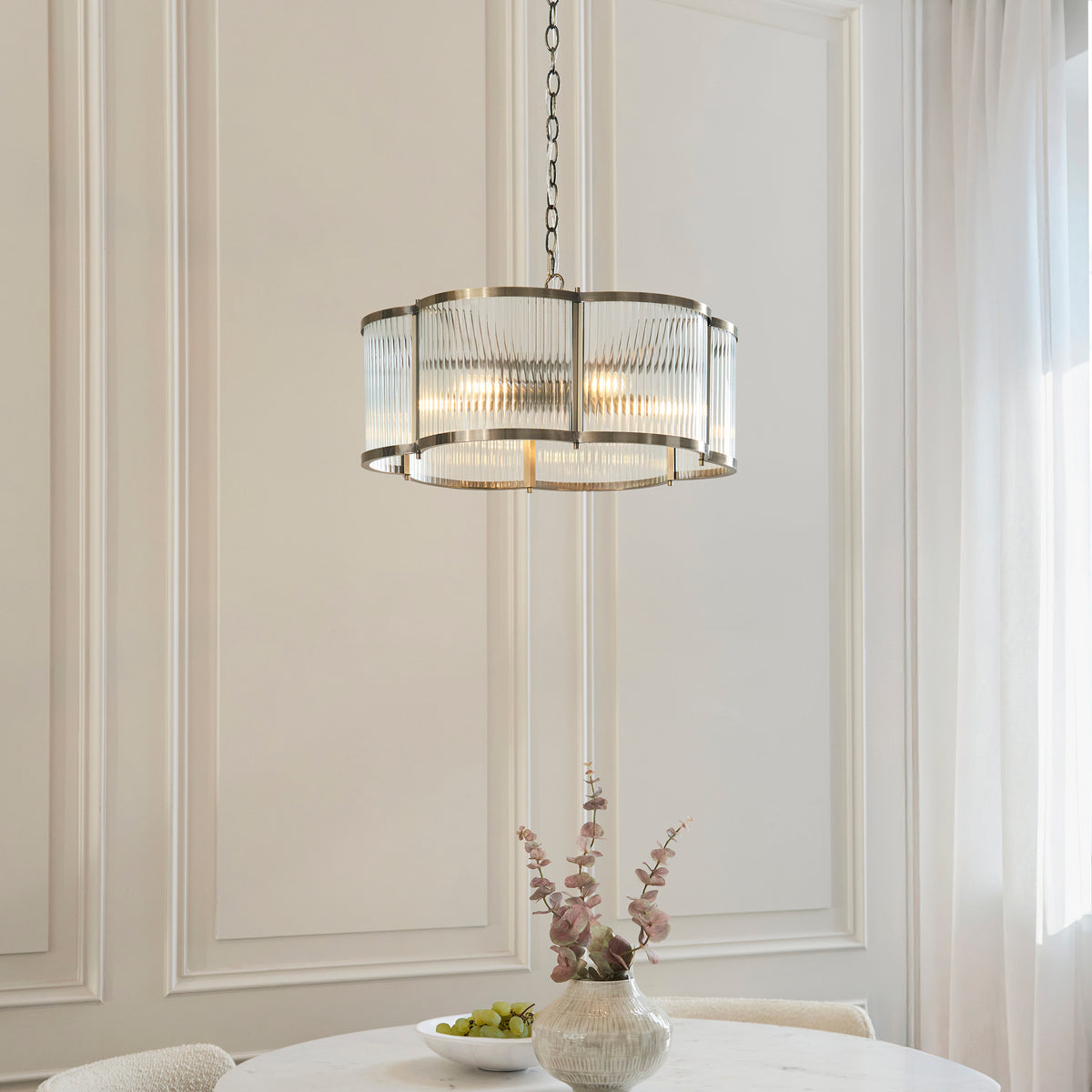 Endon Ridgeton Scallop five light pendant with clear ribbed glass panels in a brushed silver tone frame, lit warm above a round dining table with a fruit bowl and pink foliage against a white panelled wall.