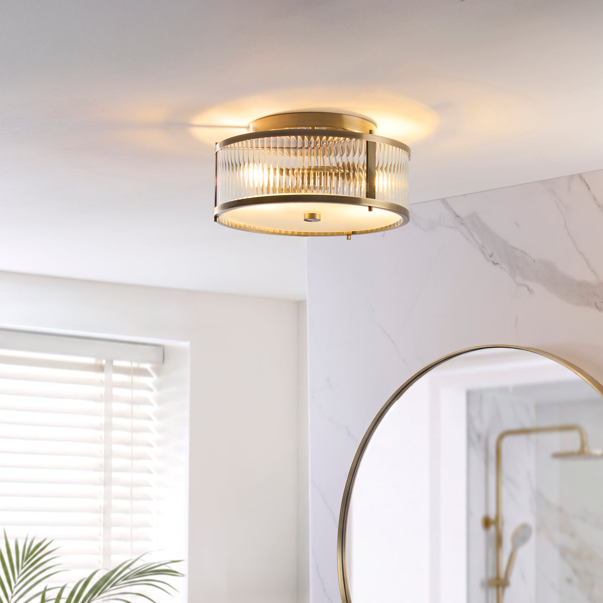 Endon Ridgeton IP44 2 light flush ceiling light in antique brass with clear ribbed glass lit in a bathroom with marble effect walls and a round brass framed mirror