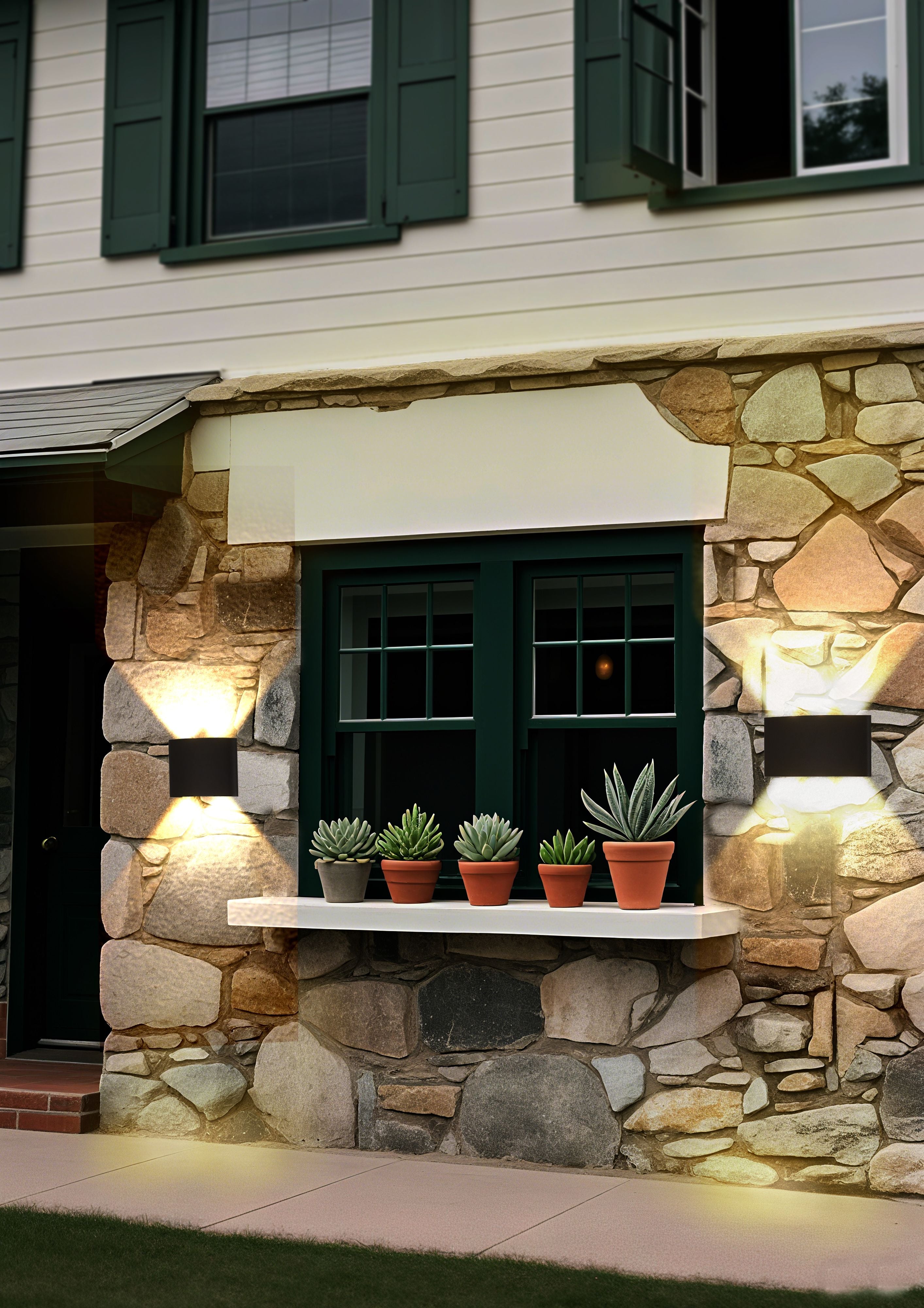 Two Shadows LED outdoor wall lights mounted either side of a window on a stone and rendered house wall, illuminated at dusk with potted succulents on the windowsill between them